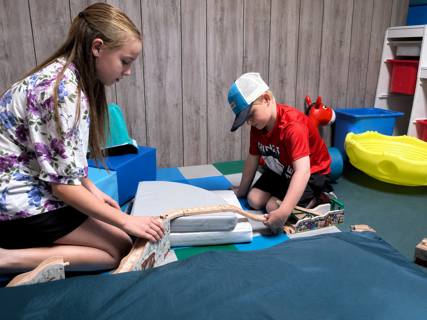 A girl and a boy playing with toy train tracks on a padded floor in a playroom. The girl has long hair and is wearing a floral shirt, while the boy is wearing a red shirt, shorts, and a cap. There are colorful toys and storage bins in the background.