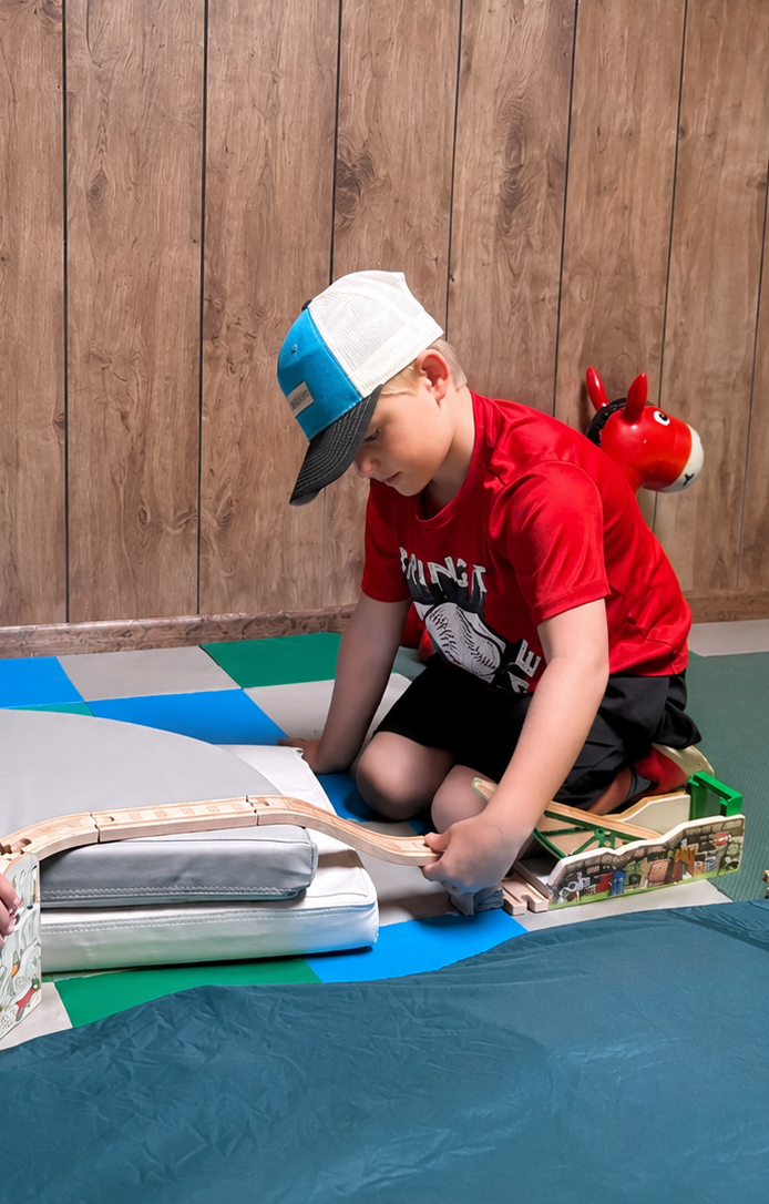 A young boy wearing a baseball cap and red t-shirt is playing with a wooden train set on a colorful foam mat against a wood-paneled wall.