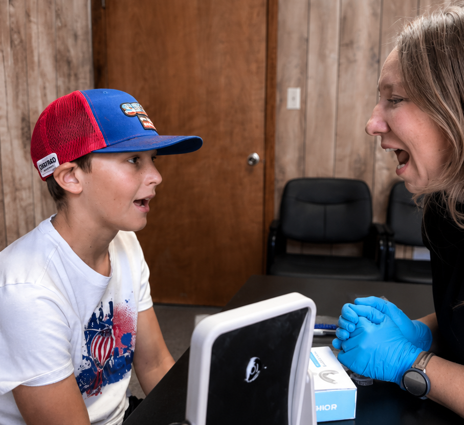 A young boy dressed casually with a red and blue baseball cap, sitting at a table with a woman wearing blue gloves and a black shirt. She appears to be a healthcare worker and is interacting with a medical device on the table.