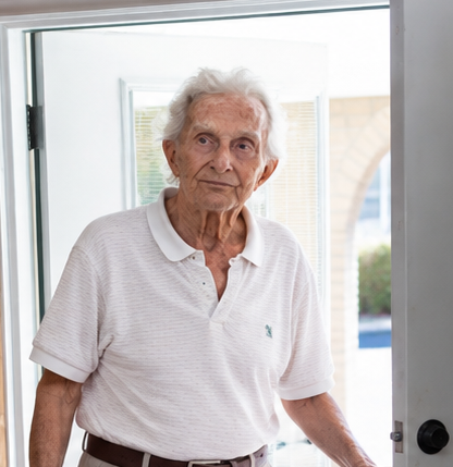 An elderly woman with white hair standing in a doorway, wearing a white polo shirt.