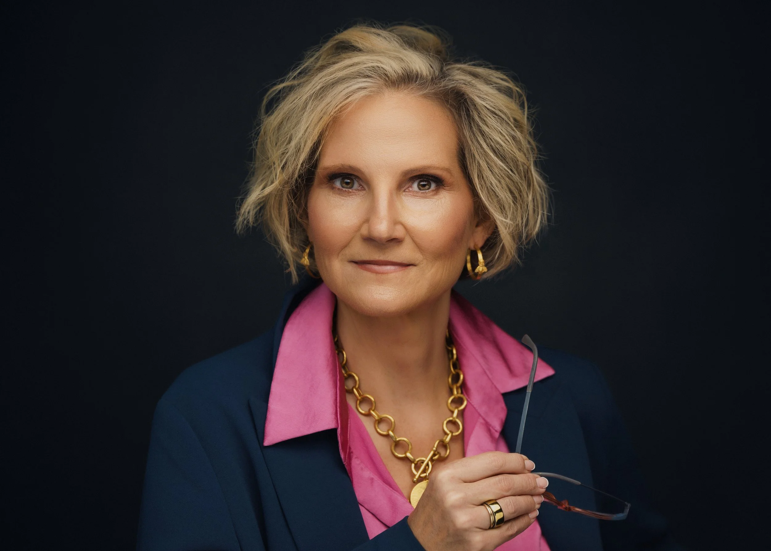 A woman with short blond hair, wearing a navy blazer, pink blouse, and gold jewelry, holding glasses and a microphone against a dark background.