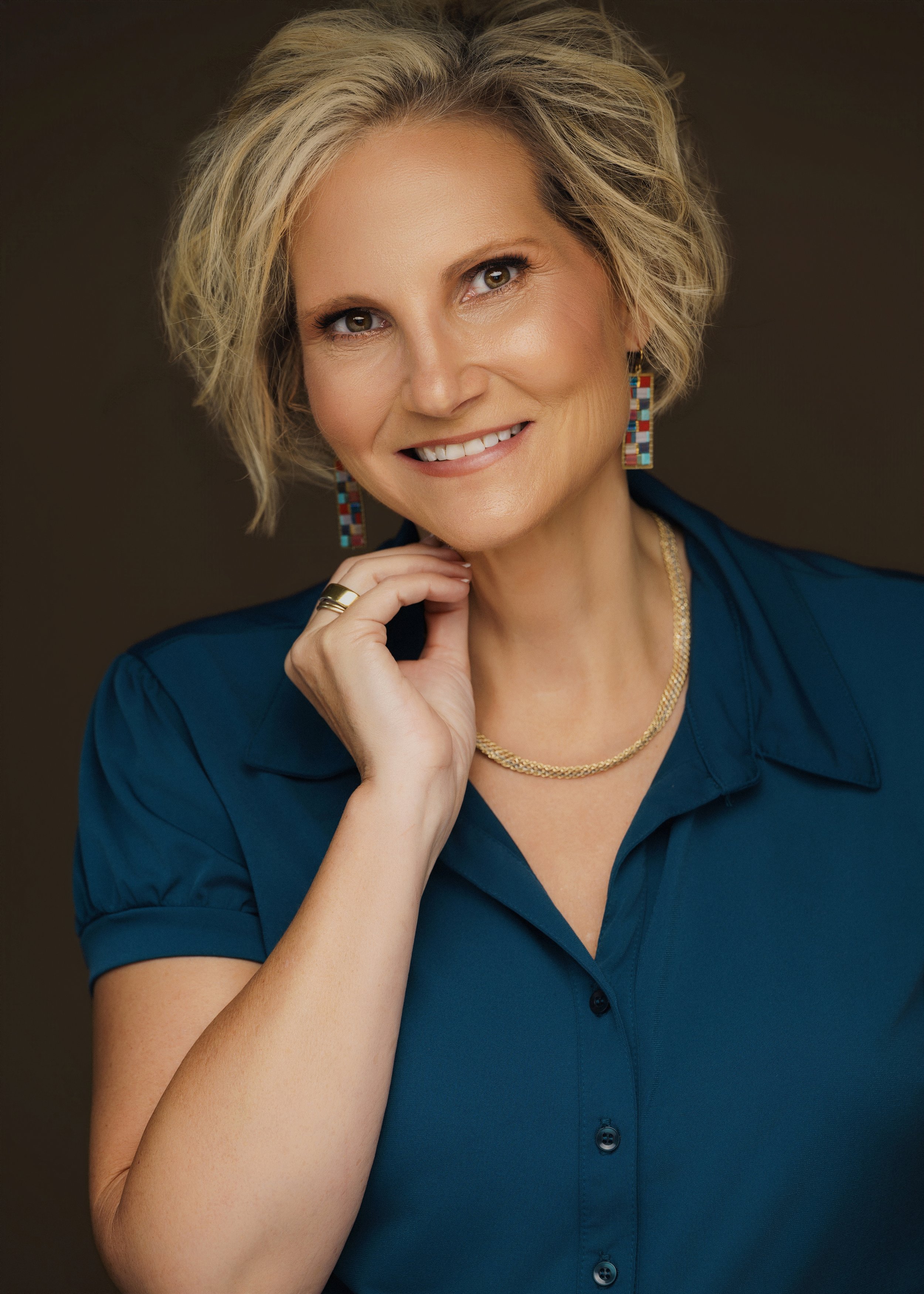 A middle-aged woman with short, wavy blonde hair, wearing a blue collared shirt, gold necklace, and colorful geometric earrings, smiling softly and resting her chin on her hand against a dark background.