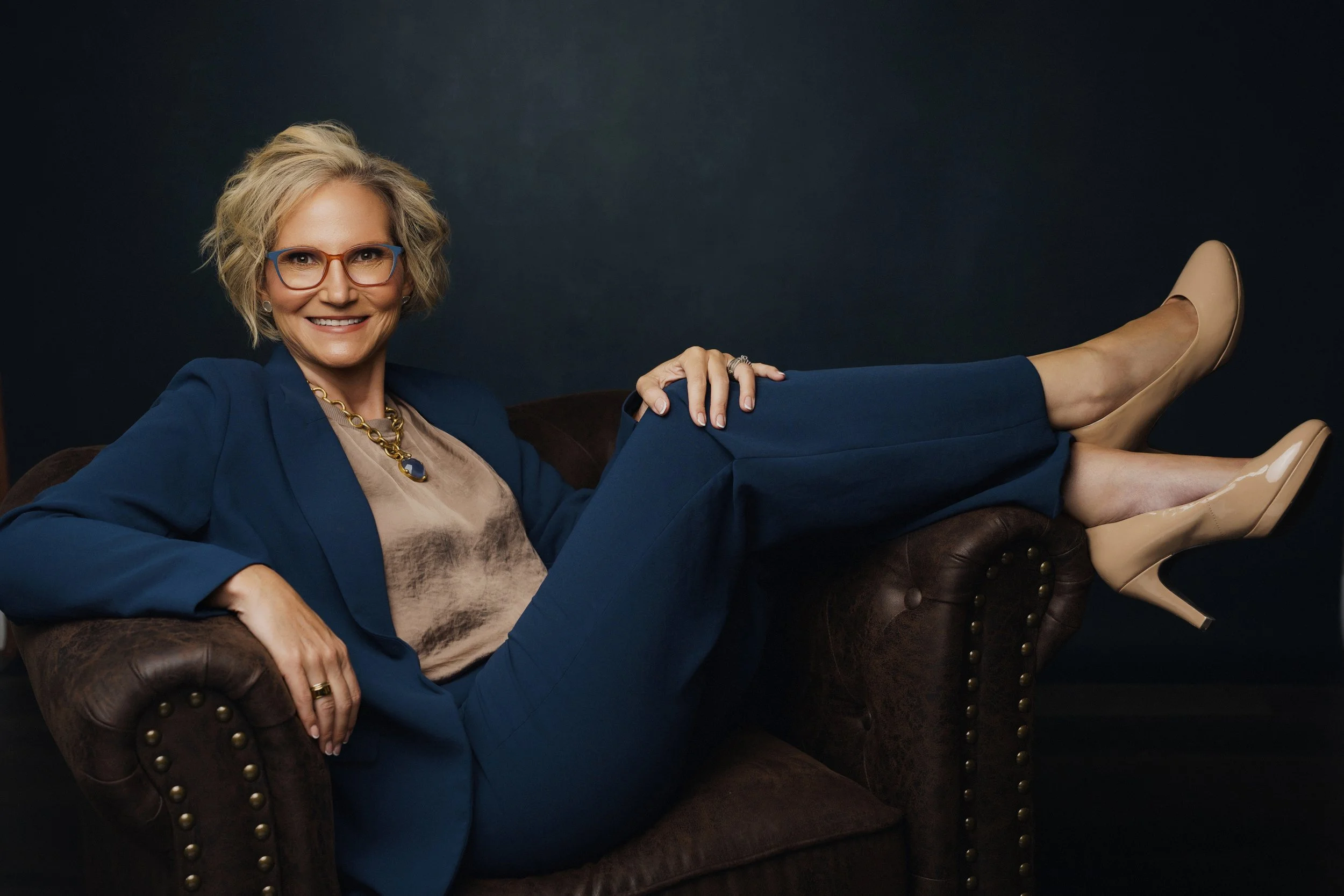 A smiling woman with short blonde hair, wearing glasses, a blue blazer, beige top, and jewelry, lies on a dark brown leather sofa against a black background, with her legs extended and crossed, wearing beige shoes.