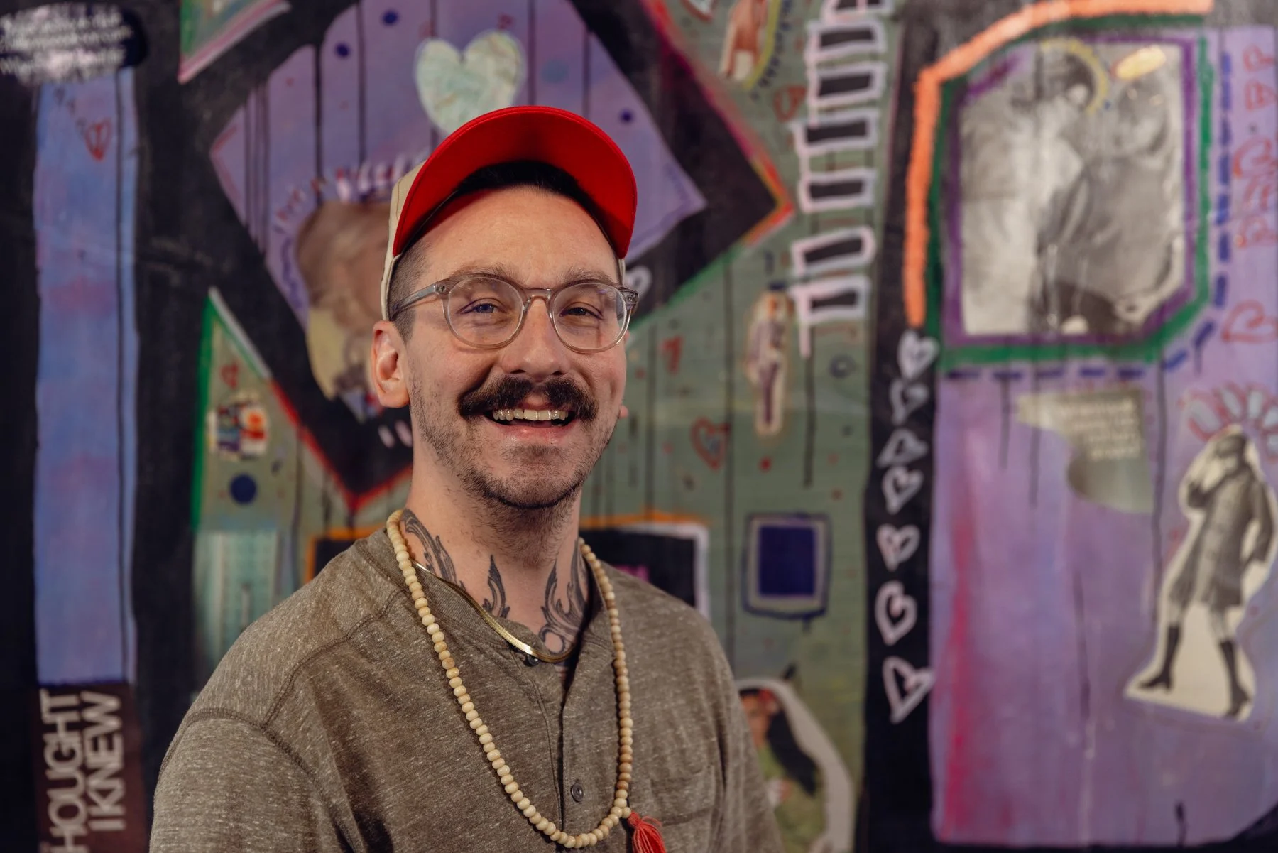 A smiling man with glasses, a mustache, tattoos on his neck, wearing a beaded necklace, a beige cap, and a brown hoodie, standing in front of a colorful mural with various drawings and text.