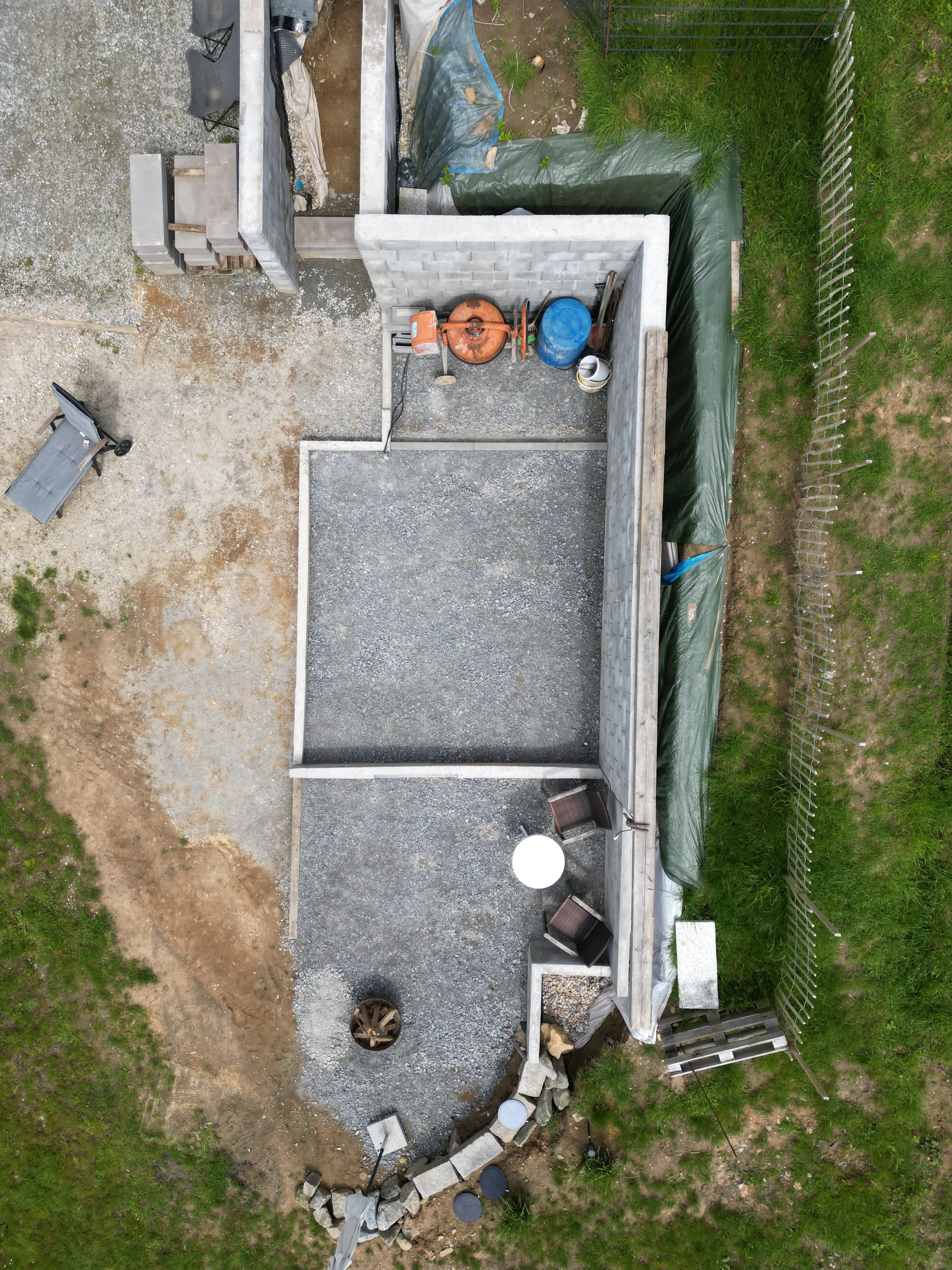 Der Bau eines Gartengebäudes mit Sicht auf eine Mauer mit Ziegeln, Bauwerkzeuge, Baumaterialien, ein Rasenbereich und eine verwitterte Gartenlaube. Der Bereich ist teilweise eingezäunt.