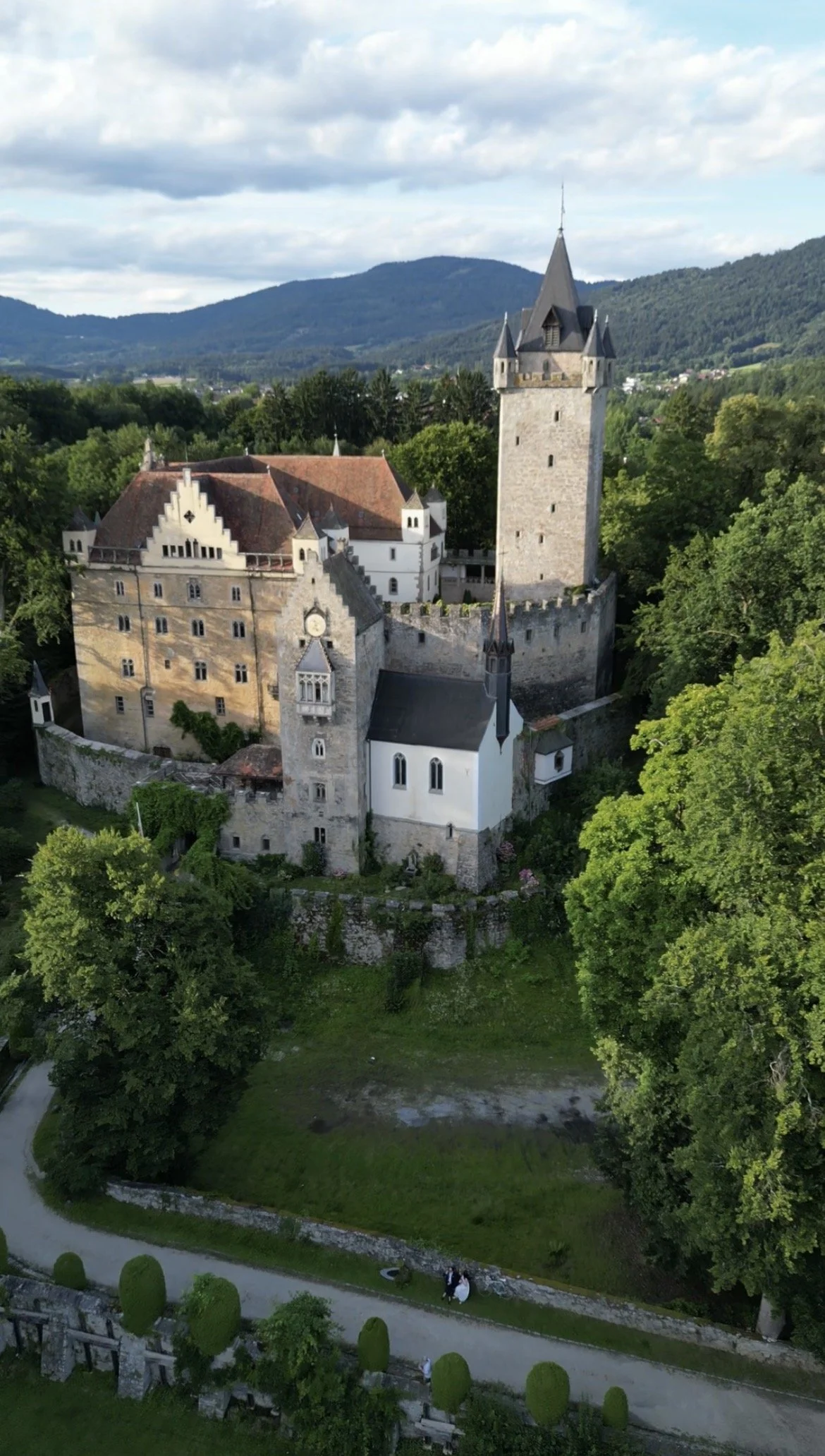 Mittelalterliche Burg auf einem Hügel, umgeben von grünen Bäumen und Wäldern, mit einem blauen Himmel und Wolken im Hintergrund.