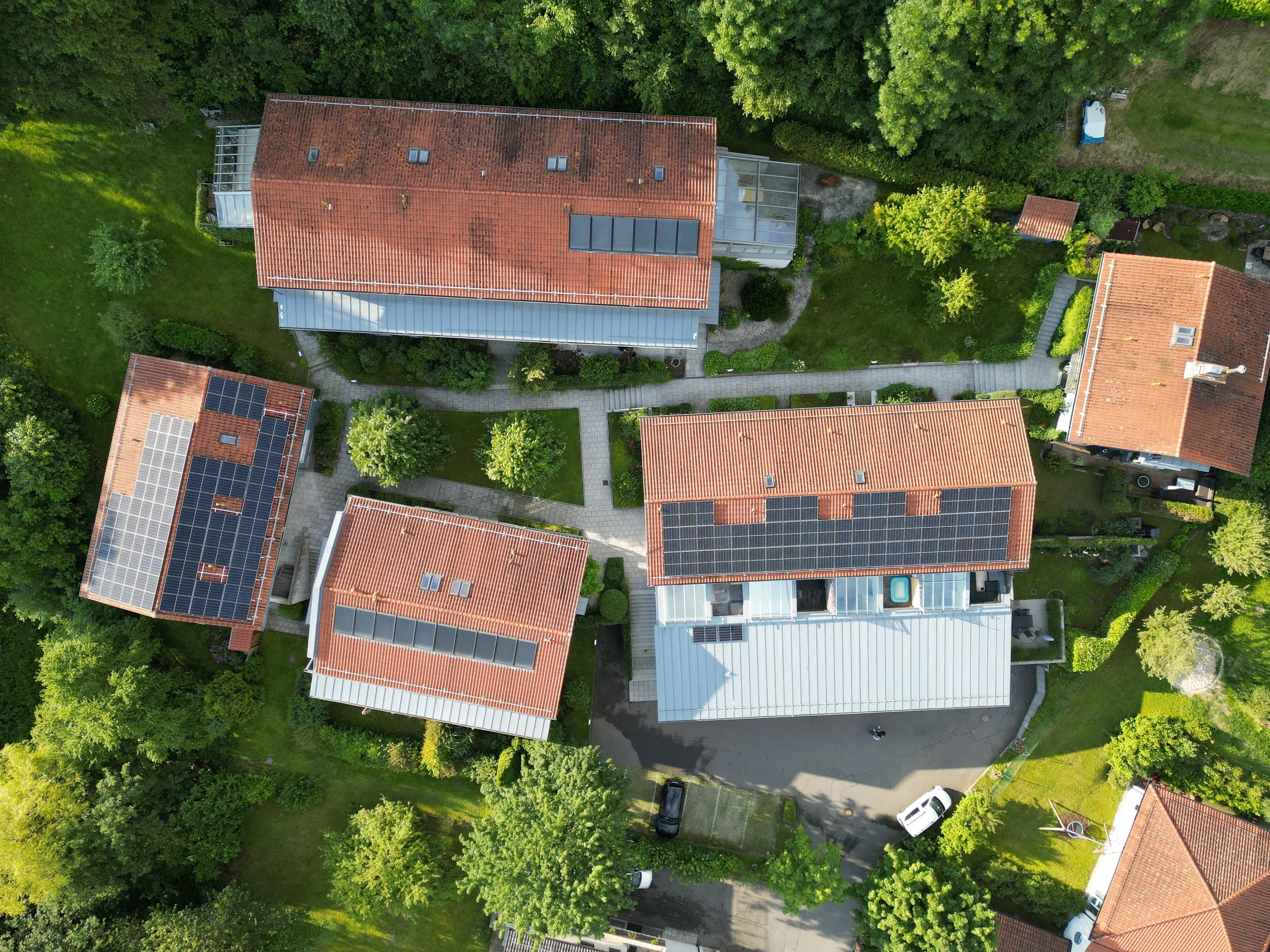 Vogelperspektive auf mehrere Häuser mit roten Dächern, einige solarbetriebene und einige mit Dachfenstern, umgeben von Bäumen, Gärten und Parkplätzen.