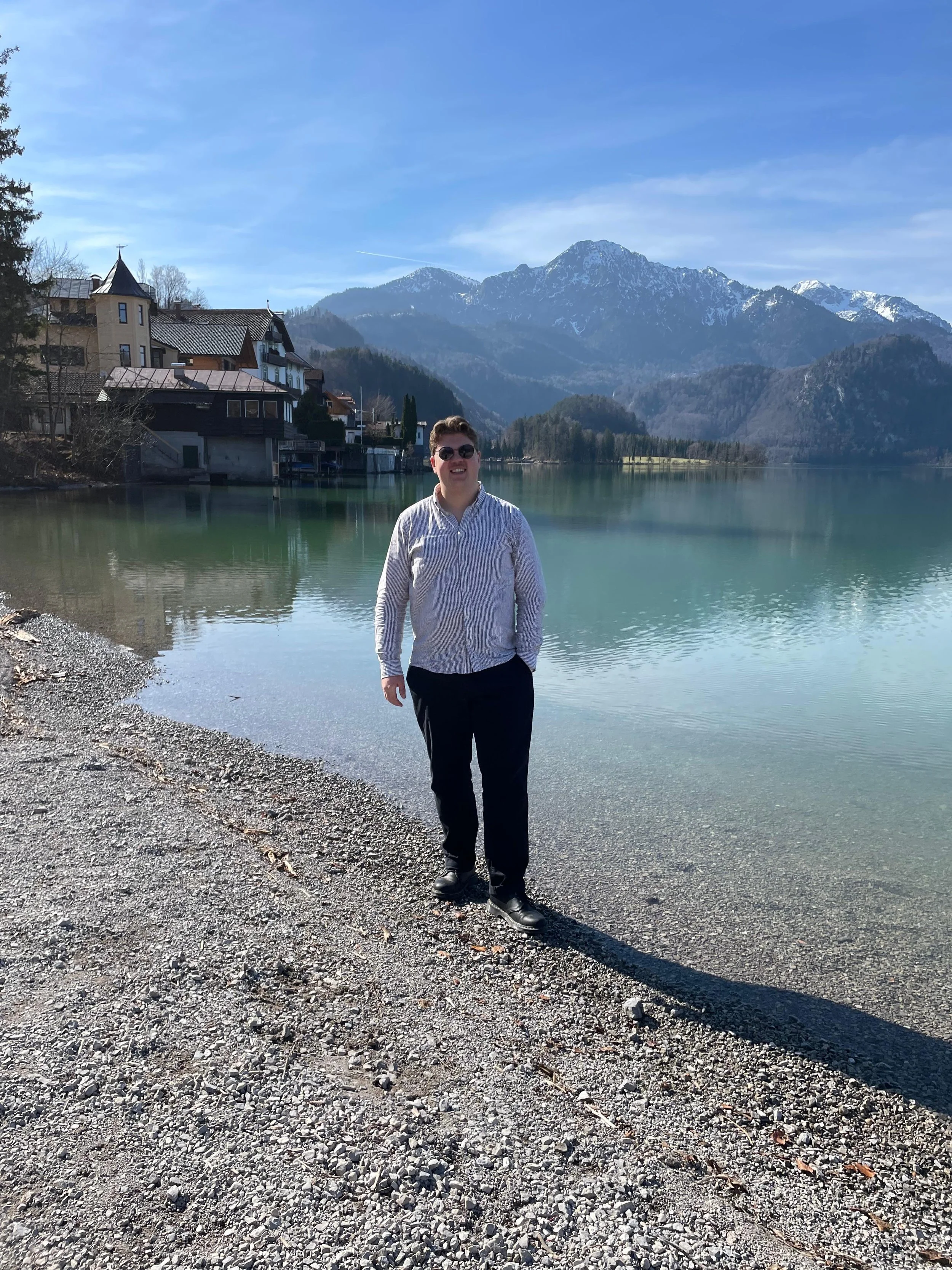 Junger Mann mit dunklen Haaren, Sonnenbrille, weißem Hemd und schwarzen Hosen steht am Ufer eines Sees mit Bergen im Hintergrund. Die Landschaft ist sonnig, der Himmel ist blau und es gibt Schneeflecken auf den Bergen.