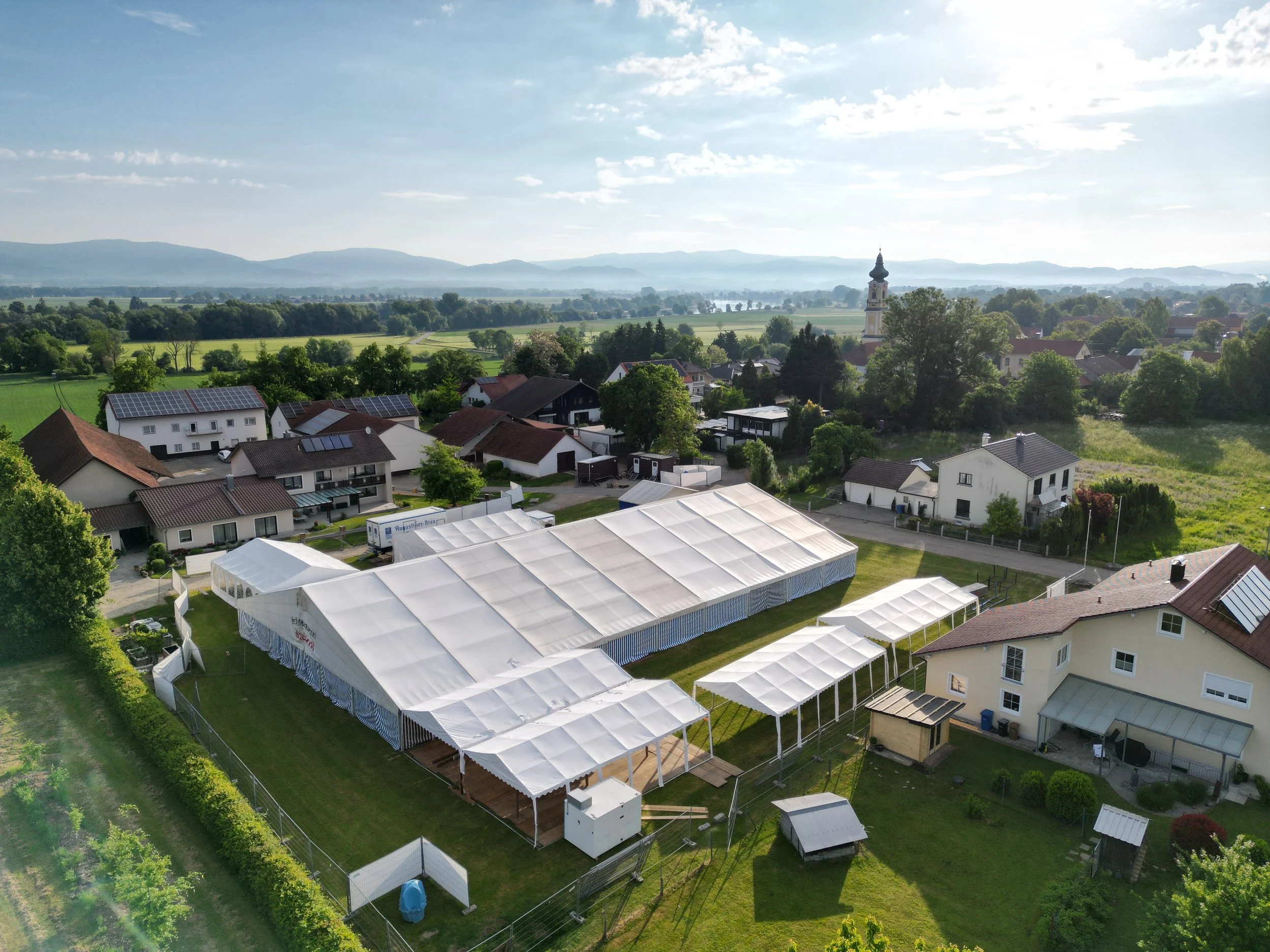 Schnappaufnahme einer ländlichen Gegend mit Wohnhäusern, großen weißen Zelten im Vordergrund, grünen Feldern und Baumwäldern in der Ferne, während die Wolken den Himmel bedecken und die Berge im Hintergrund sichtbar sind.