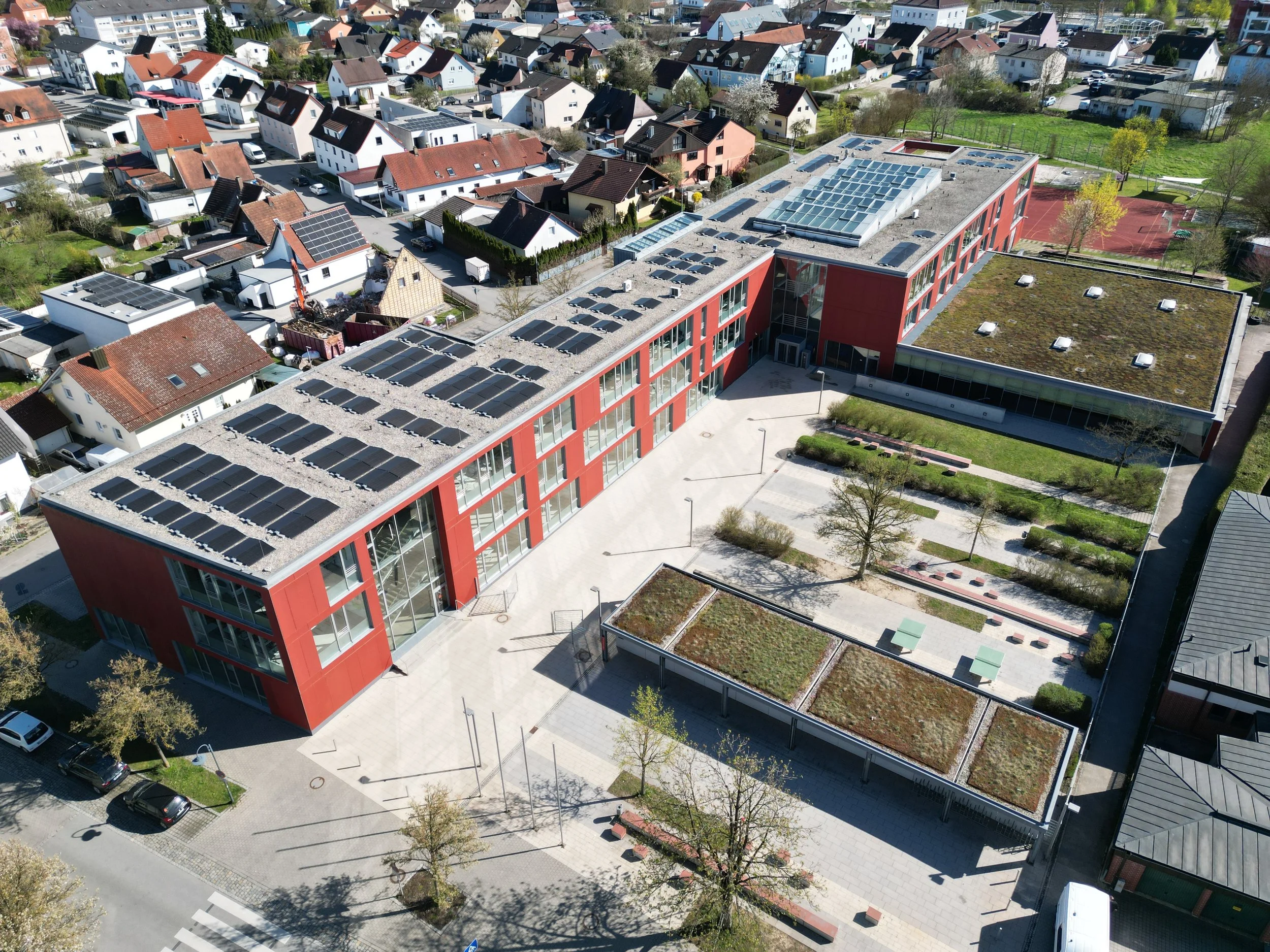 Luftaufnahme eines Schulgebäudes mit grünem Dach und umgebenden Schulhof sowie einem Nachbarschaftsviertel mit Häusern und Straßen.