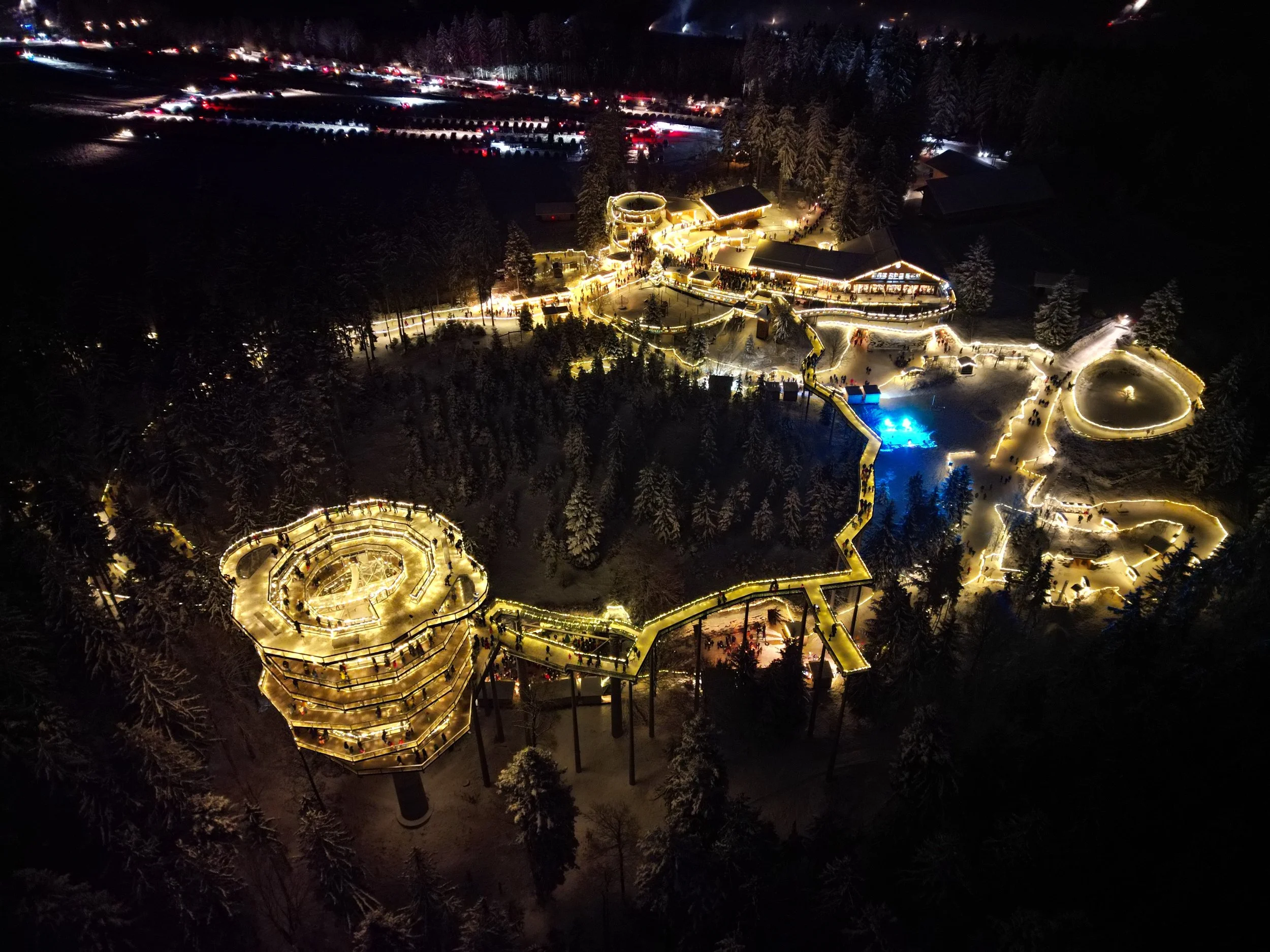 Nachtaufnahme eines beleuchteten Weihnachtsmarktes im Schnee mit Rutschen und festlich geschmückten Gebäuden in einem Waldgebiet.