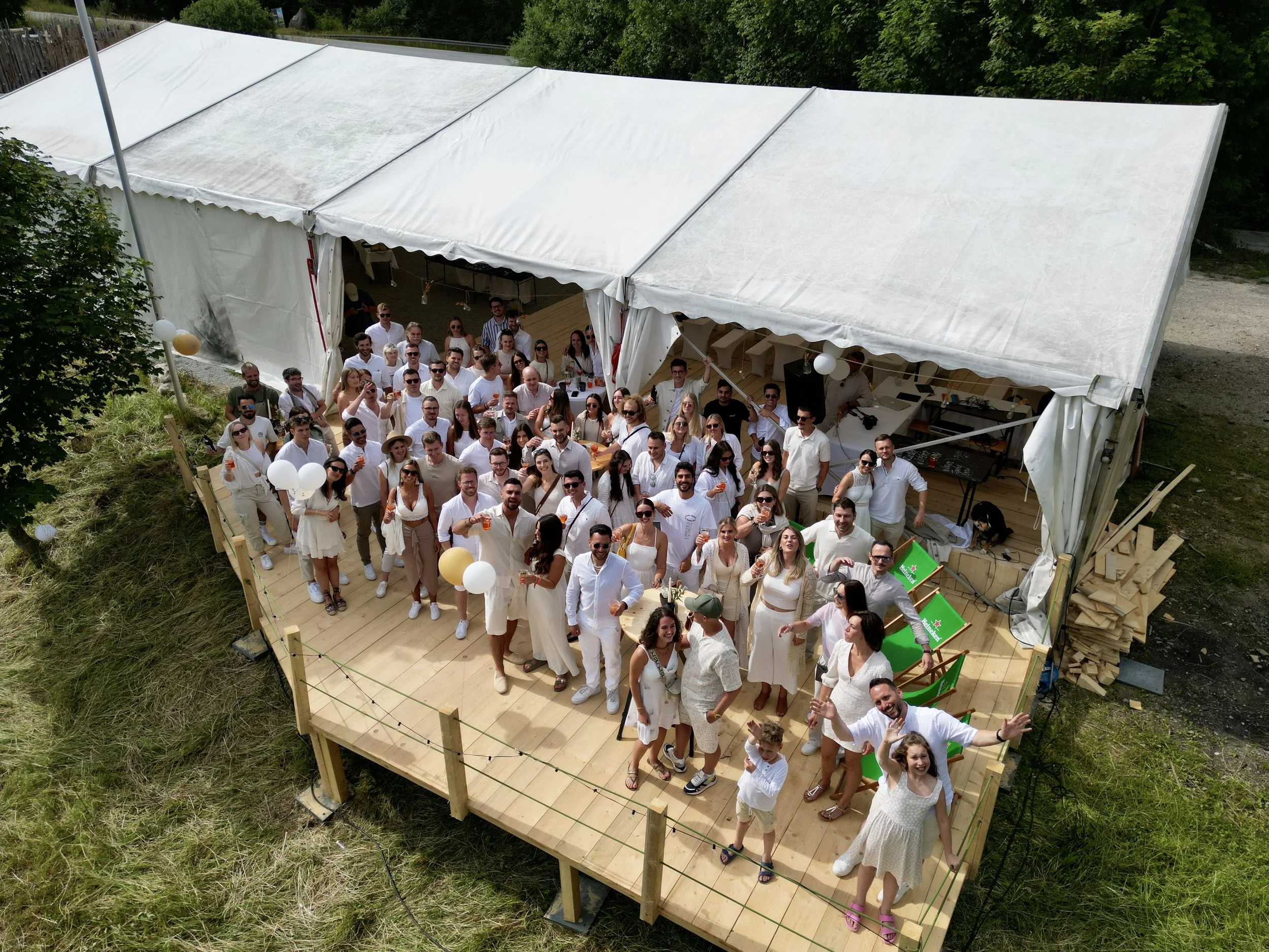 Eine große Gruppe von Menschen feiert auf einer hölzernen Terrasse unter einem weißen Zeltdach im Freien, umgeben von Bäumen und Grünflächen, bei einer Party oder Veranstaltung.