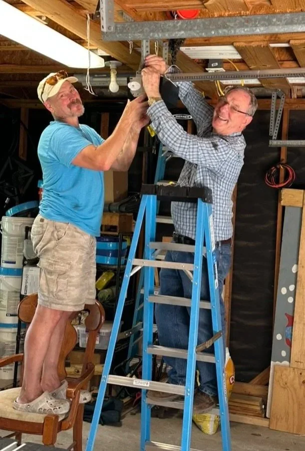 Two men working together to install a garage door opener, with one standing on a chair and the other on a ladder, both smiling.