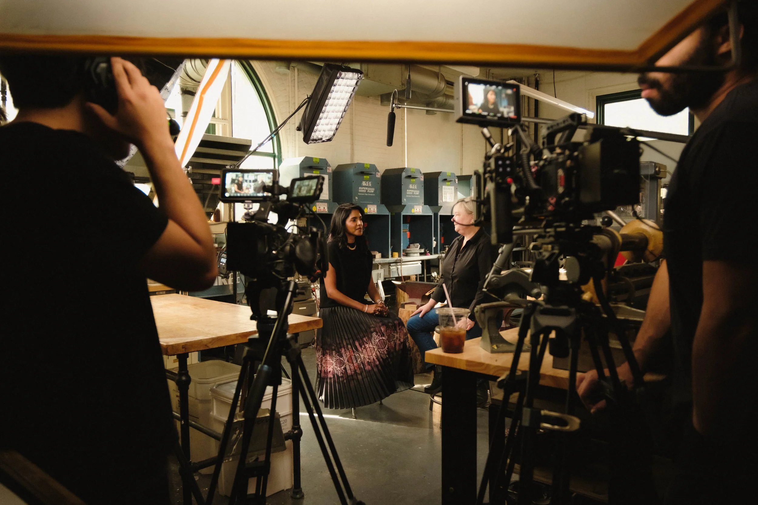 Behind-the-scenes of a video or film shoot with cameras and crew capturing two women talking, one with dark hair and dressed in black, another with white hair and glasses, in an industrial kitchen setting with machinery and kitchen equipment.