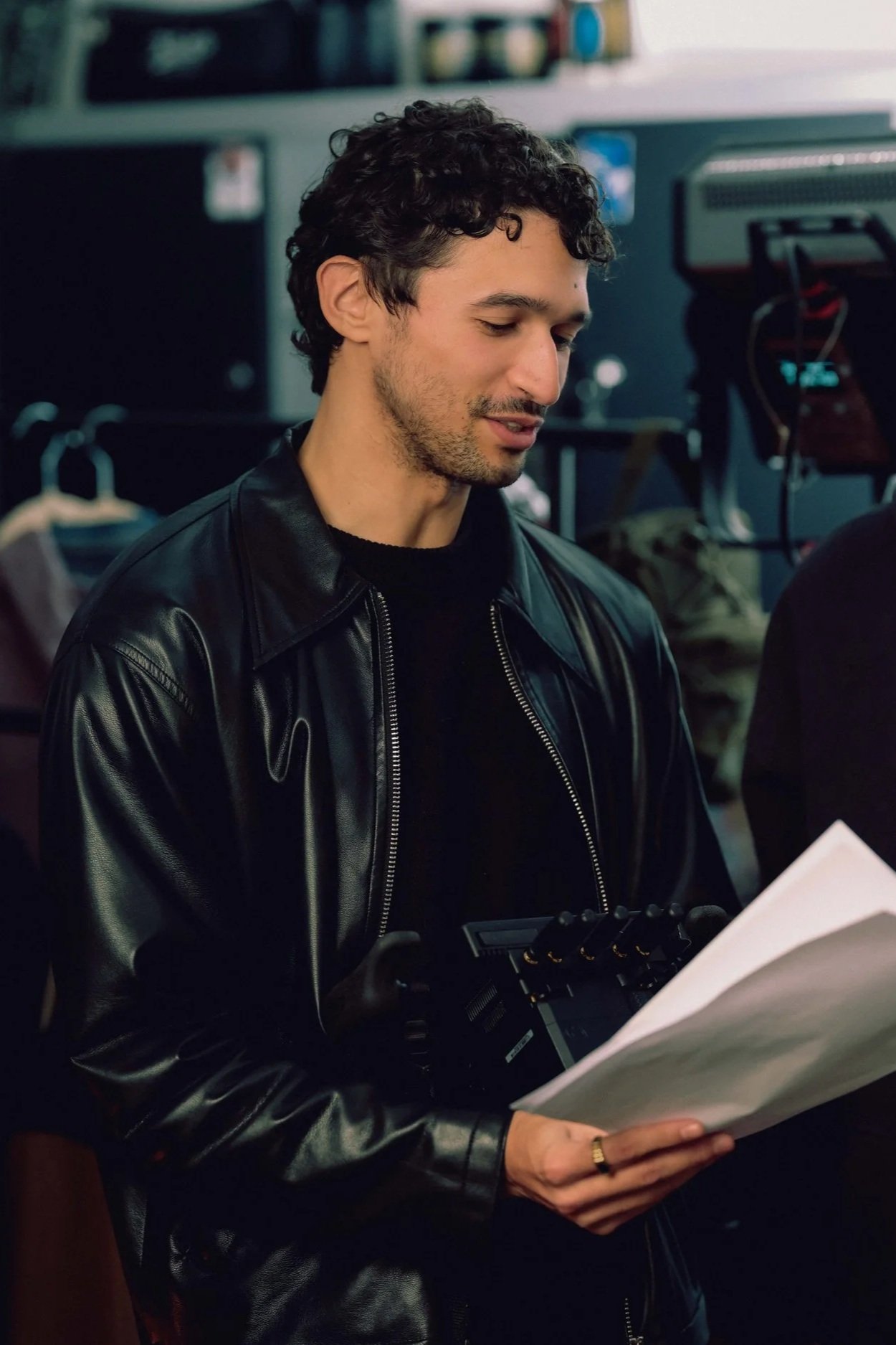 A young man with curly dark hair and light skin is wearing a black leather jacket and holding a piece of paper and a device while looking at the paper in an indoor setting, possibly a store or workspace.