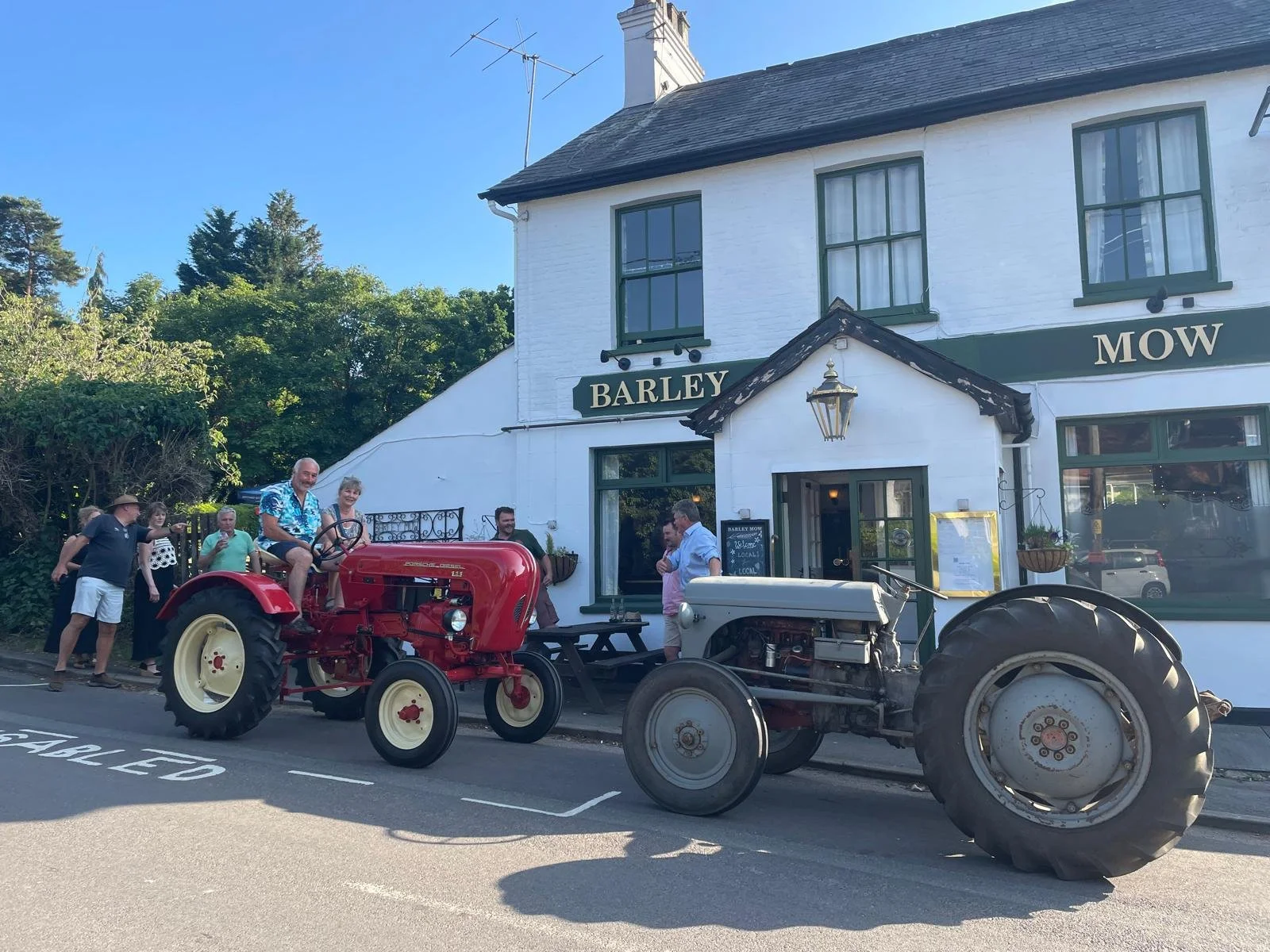 A group of people gathered outside a white building with green trim that has signs reading "Barley" and "Mow." Two vintage tractors, one red and one white, are parked in front. Some people are standing and talking, while one person is sitting on the 