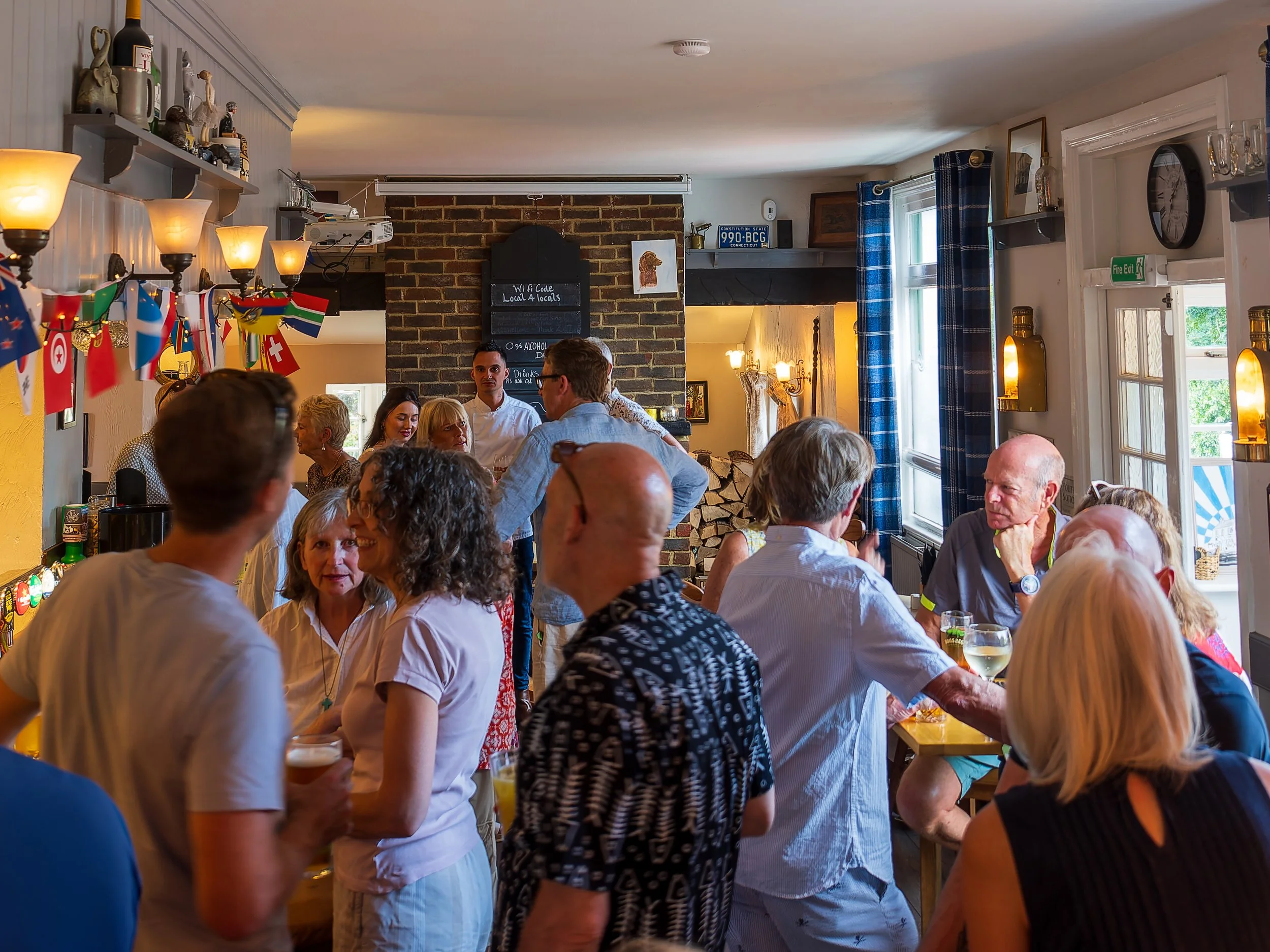 People gather in a cozy restaurant or pub, socializing and enjoying drinks, with a decor featuring flags, a brick chimney, and warm lighting.