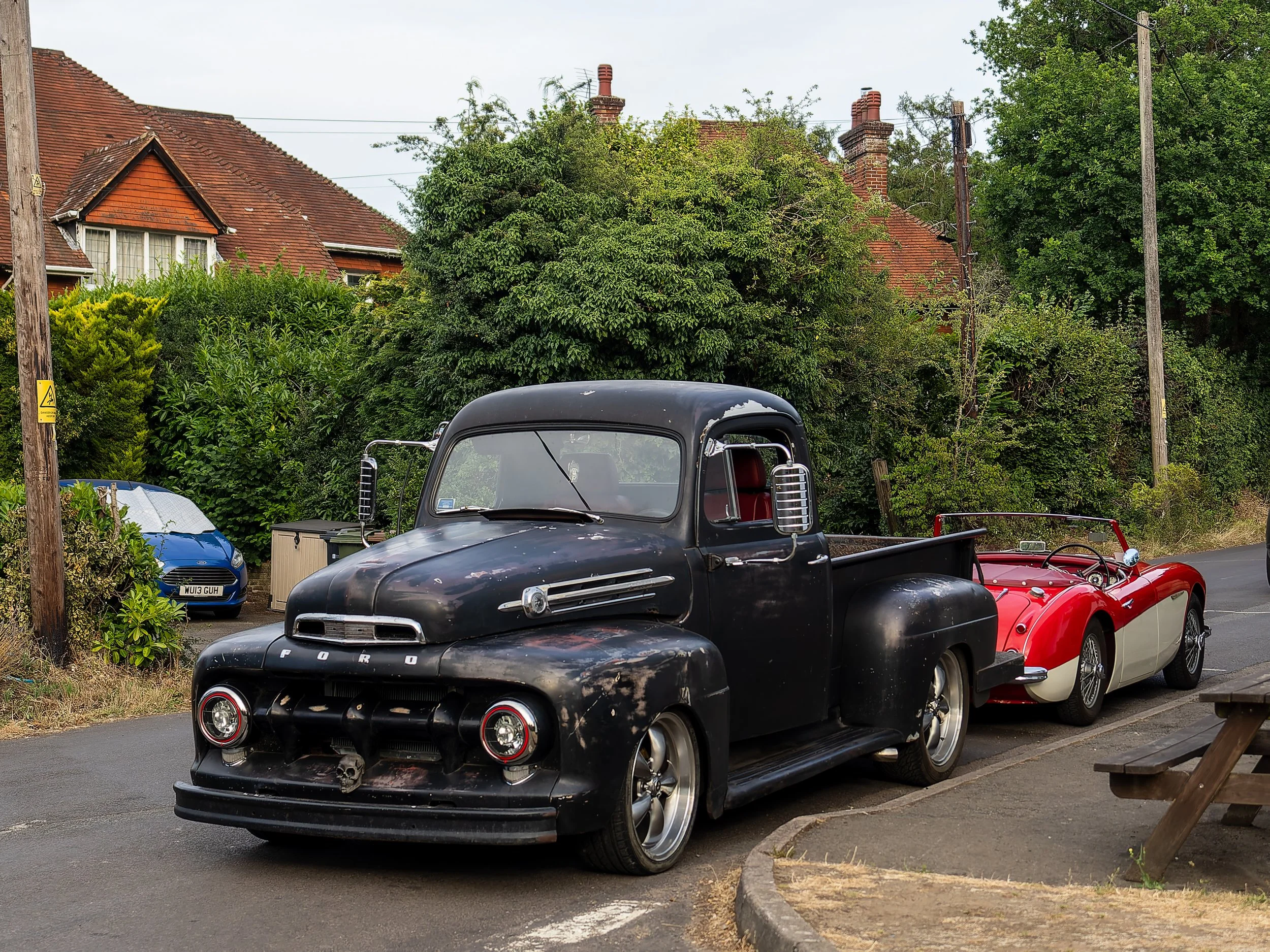 A row of vintage cars parked along a residential street, with houses and greenery in the background.