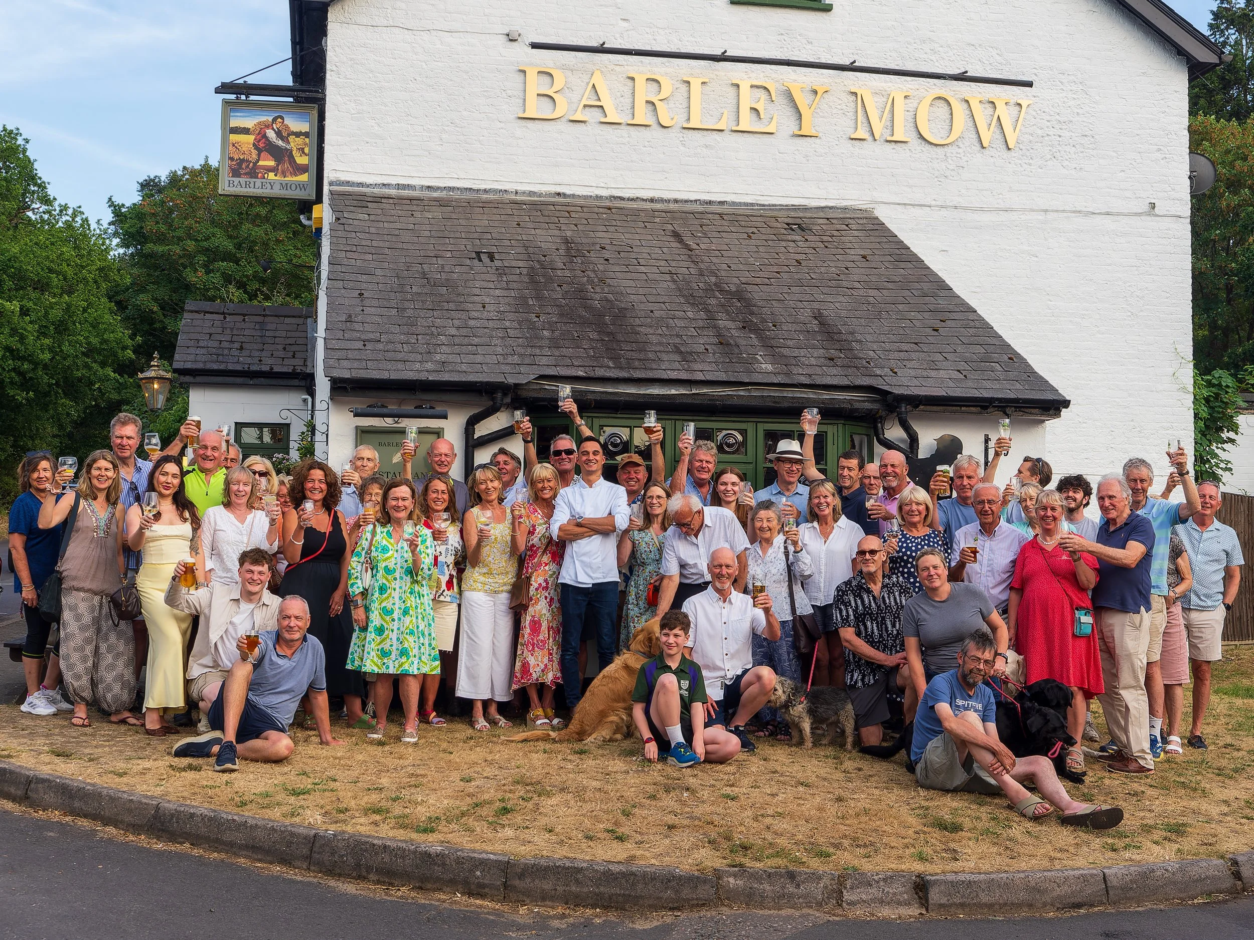 A large group of people gathered outdoors in front of a white building with a sign that reads 'Barley Mow', many holding up glasses of beer and smiling, suggesting a celebration or community event.