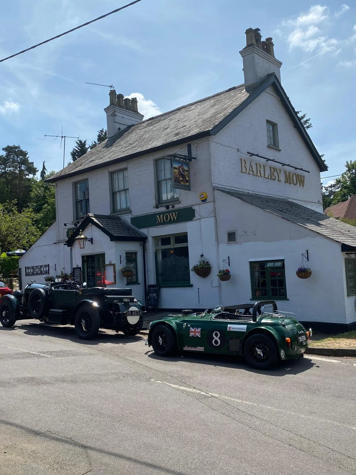 The image shows a white, two-story pub building called 'Barley Mow' with a traditional style. There are hanging flower baskets outside, and a couple of vintage cars parked in front: a black one and a green one with racing decals and the Union Jack. T