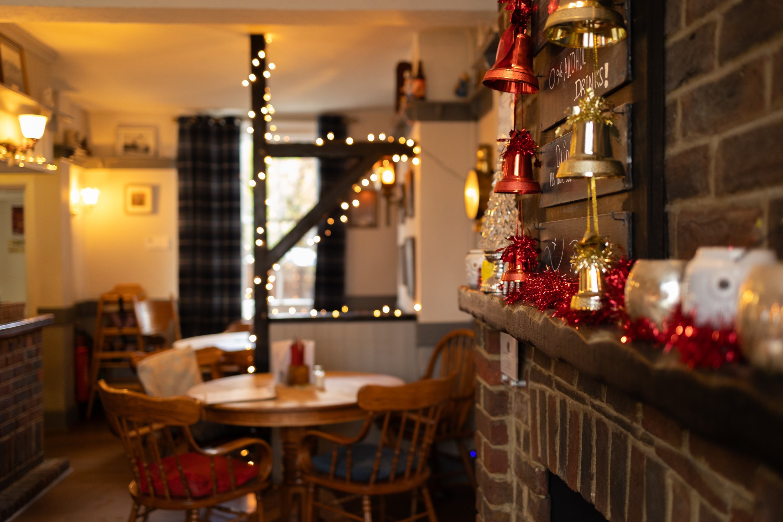 A cozy restaurant interior decorated with Christmas ornaments, including red and gold tinsel, bells, and small Christmas trees on a wooden shelf mounted on a brick wall.