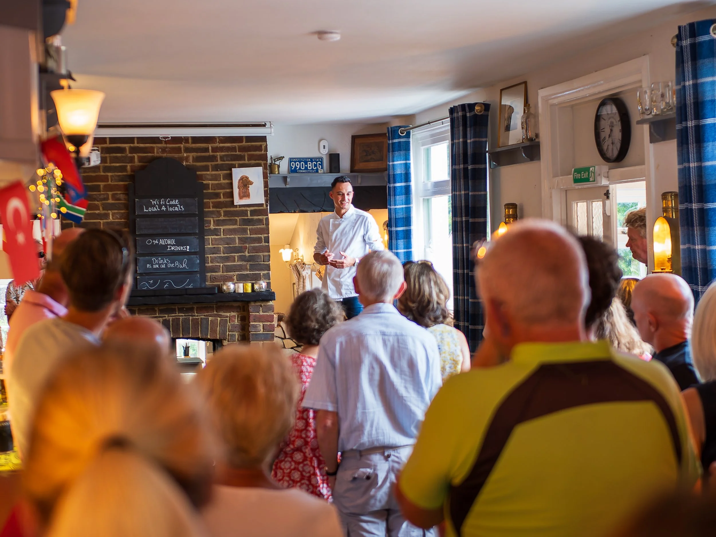 A man dressed as a chef speaking to an audience in a cozy restaurant with exposed brick walls, blue checkered curtains, and various decorations, including a blackboard with Wi-Fi code and a Connecticut license plate.