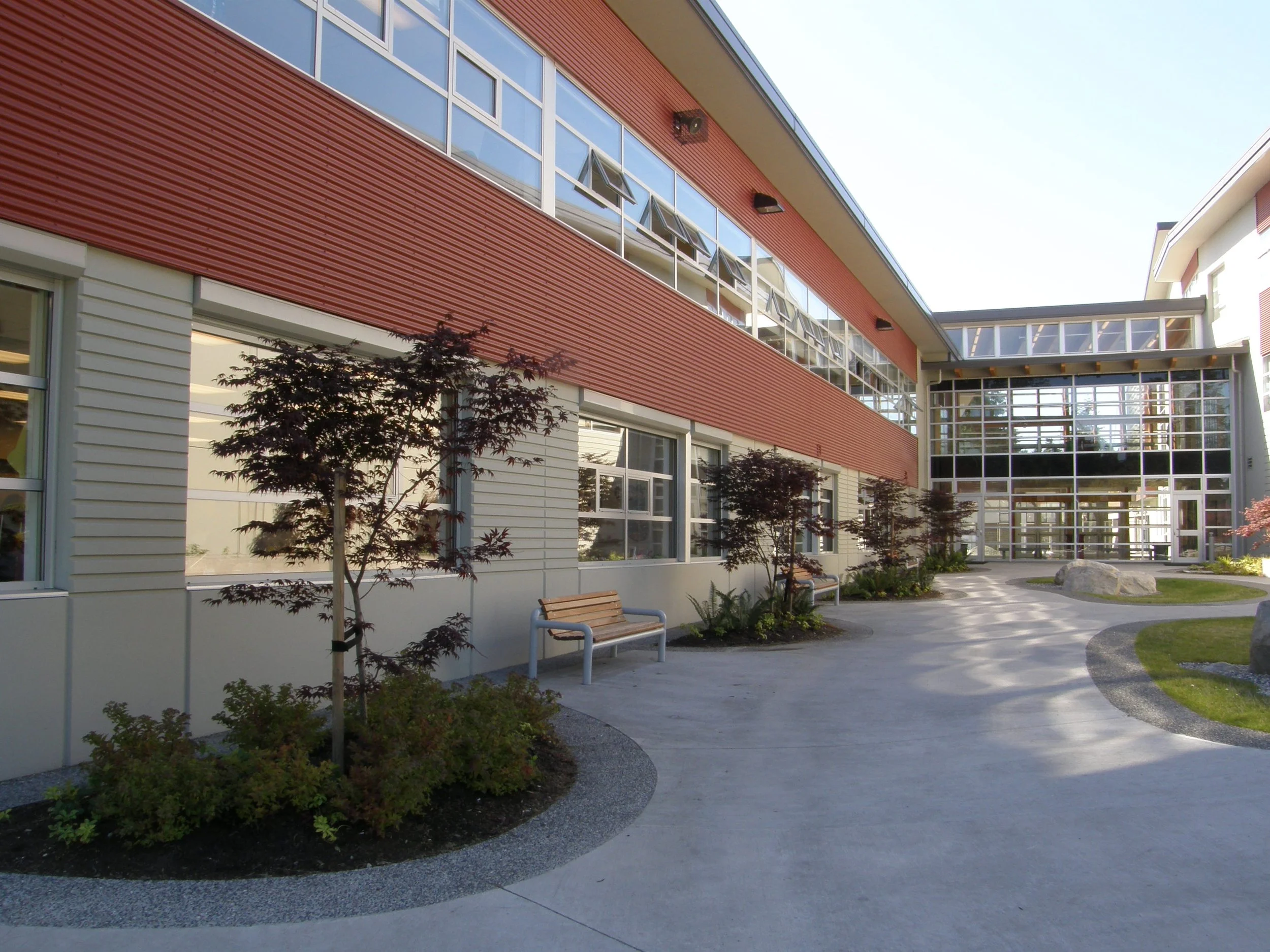 Modern building with red and white exterior, large glass windows, and a walkway with benches and small trees in the landscaped area.