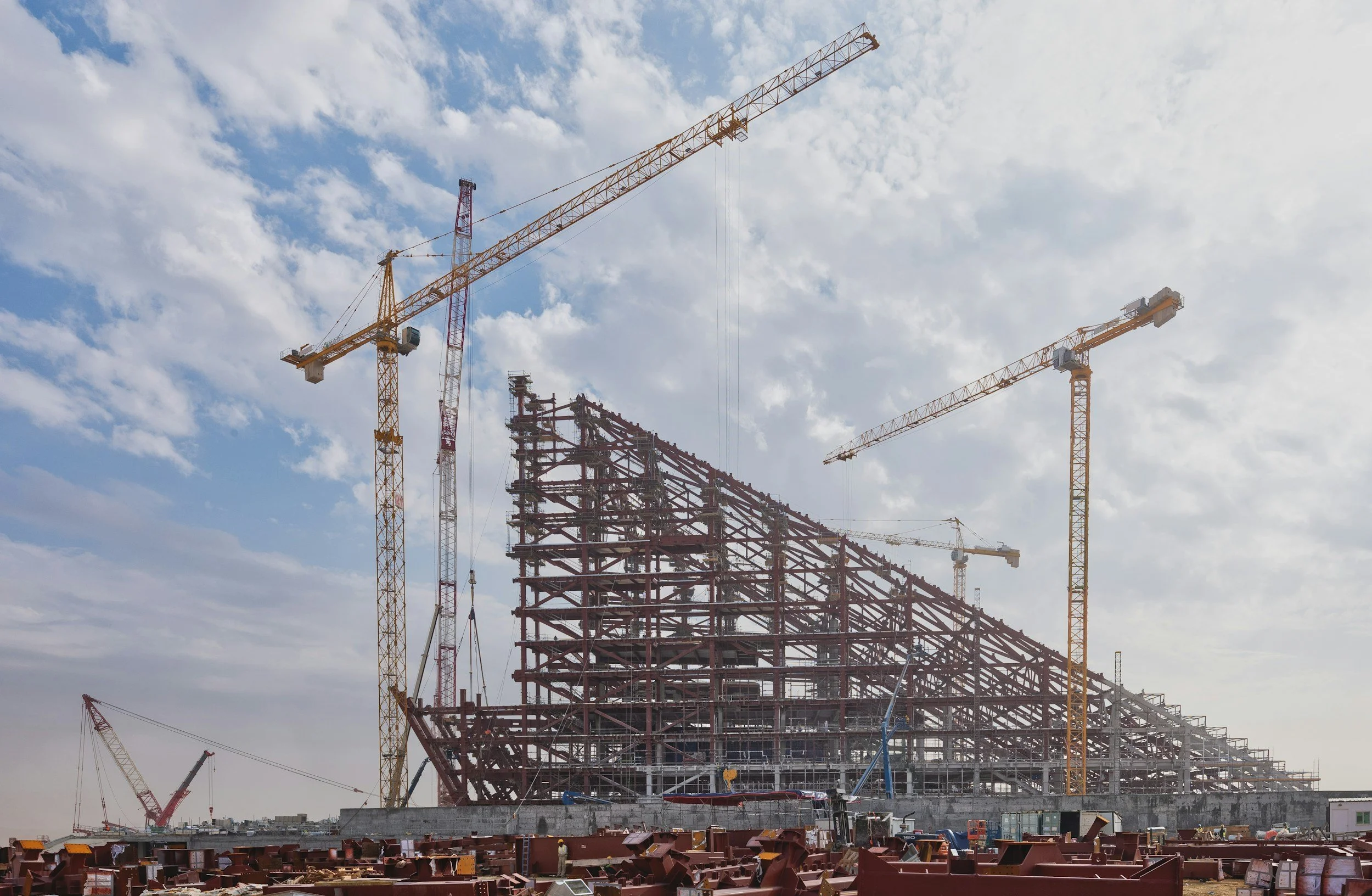 Construction site with multiple cranes and a steel framework of a building under construction against a cloudy sky.
