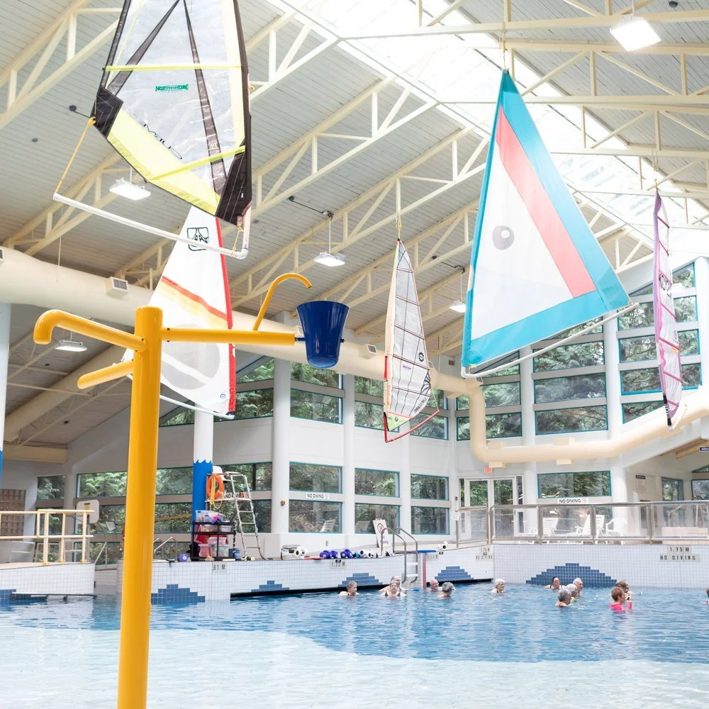 Indoor swimming pool with large windows, colorful sailboats hanging from the ceiling, and people swimming.