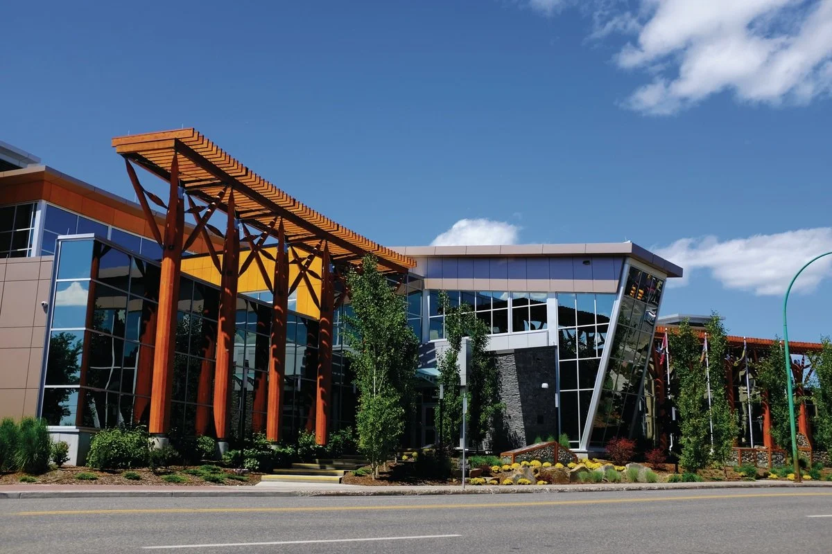 Modern building with large glass windows, wood architectural details, and surrounded by landscaping with trees and shrubs under a blue sky.