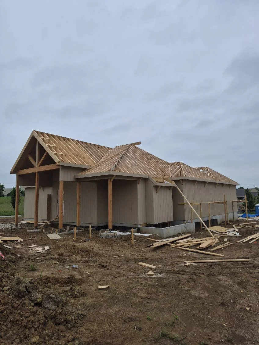 Under construction house with wooden framework and partially completed roof, surrounded by construction debris on dirt ground.