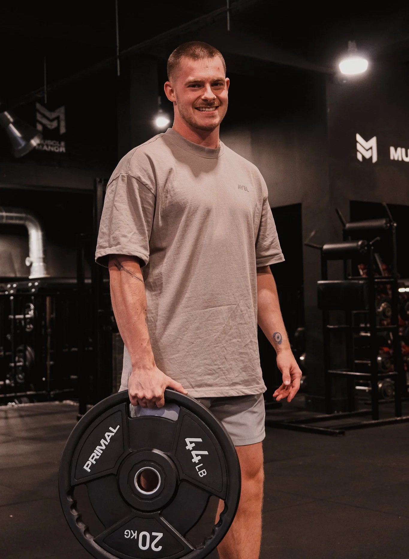 A young man with short hair and a mustache smiling in a gym, holding a 20-pound weight plate in one hand, wearing a gray t-shirt and shorts, with gym equipment and black walls in the background.