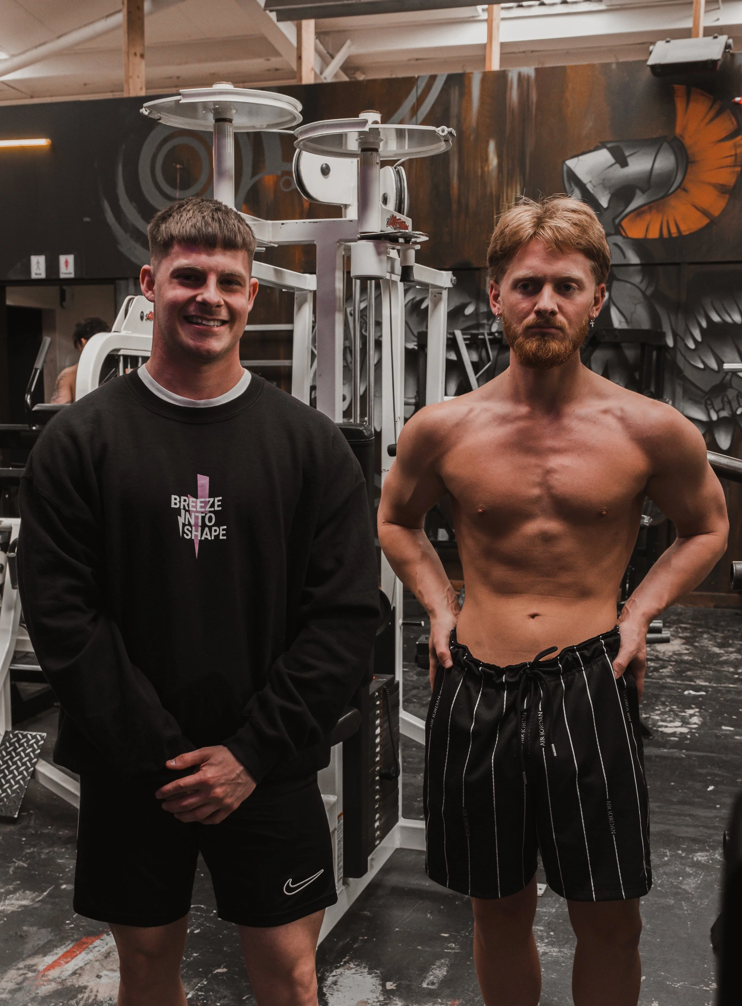 Two men standing shirtless and wearing black shorts in a gym, with various exercise equipment in the background. One man is smiling and the other has a serious expression.