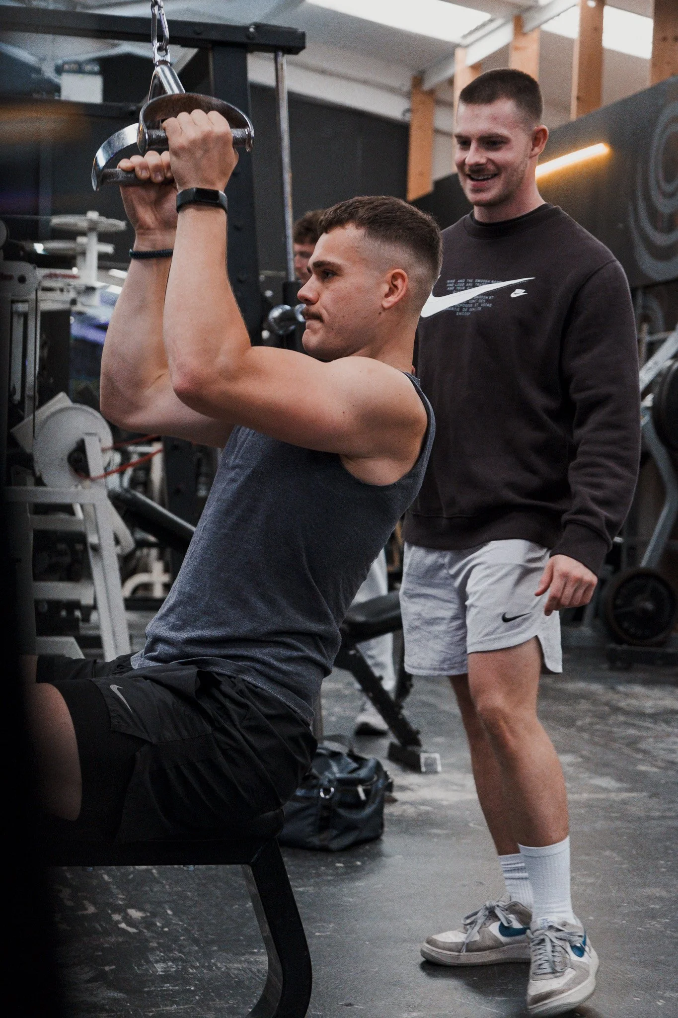 Two young men in a gym. One man is sitting on a rowing machine, pulling the handle towards his chest, while the other man stands beside him, smiling and observing. The gym has black equipment and wooden beams.