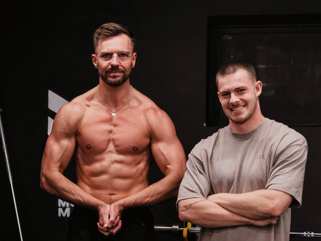 Two men posing inside weight room, one shirtless showing muscular physique, the other wearing a gray T-shirt smiling with arms crossed.