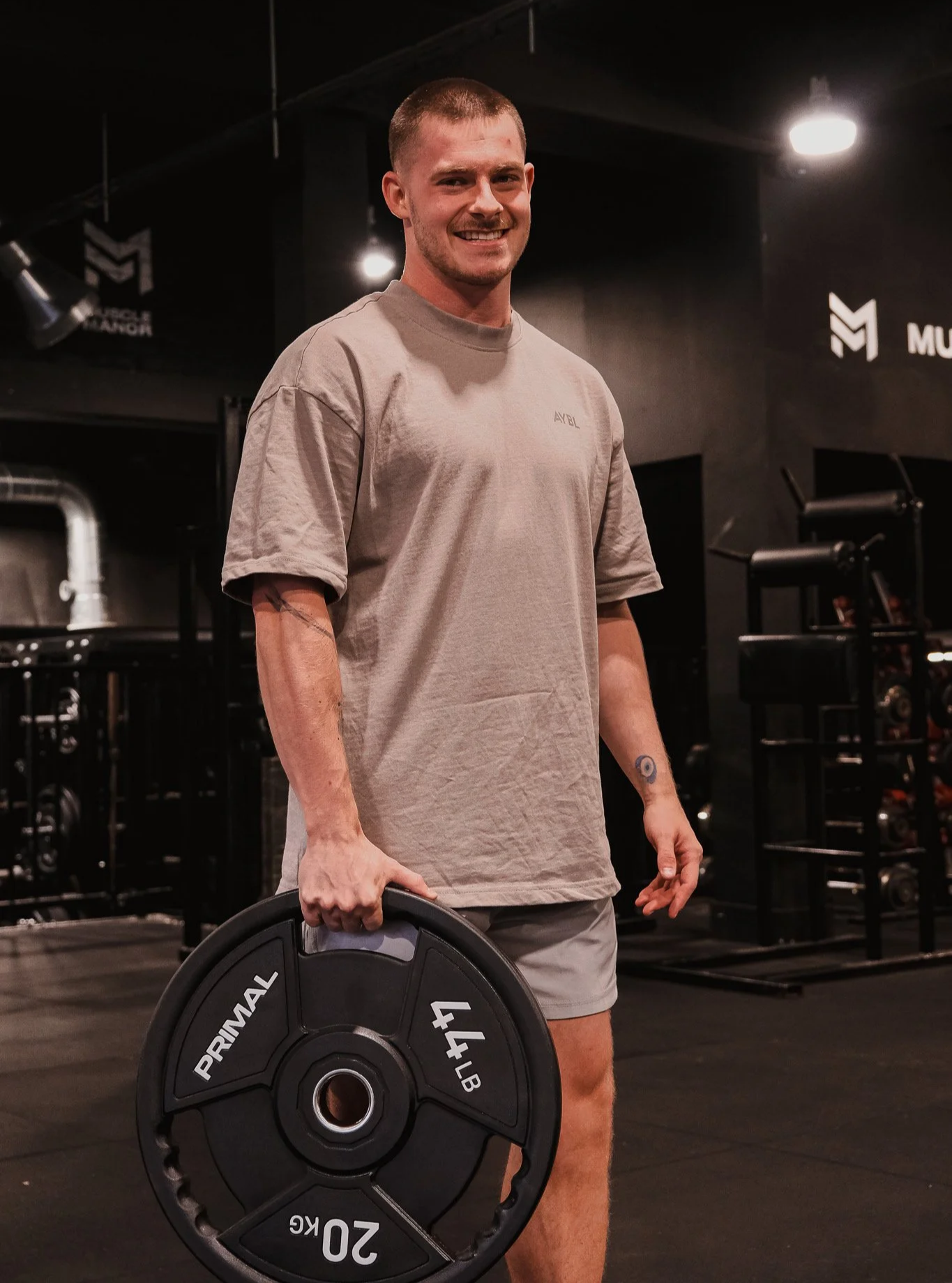 A smiling man in a beige t-shirt holding a 20 kg weight plate in a gym.