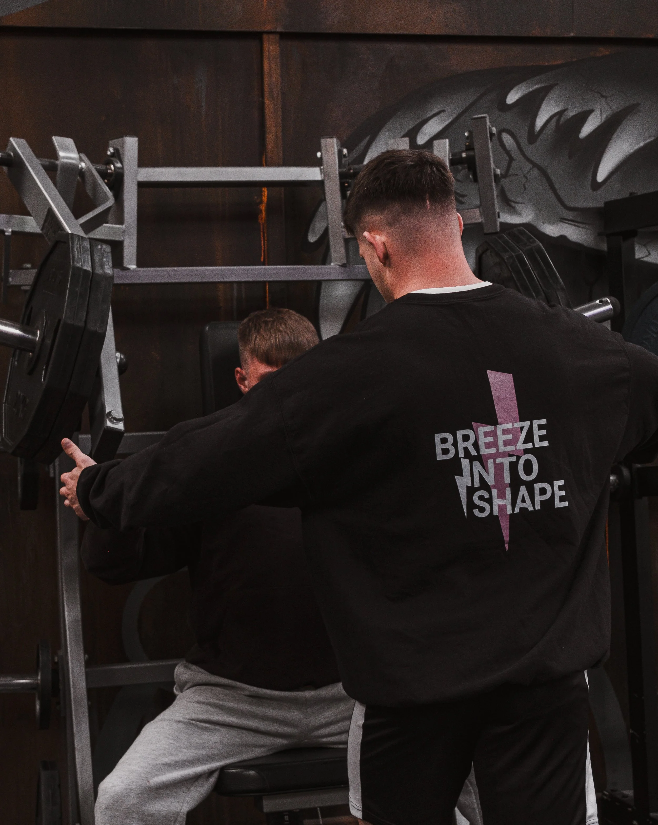 Two men sitting in a gym, one in a black hoodie with text on the back and the other in grey sweatpants, with gym equipment in the background.