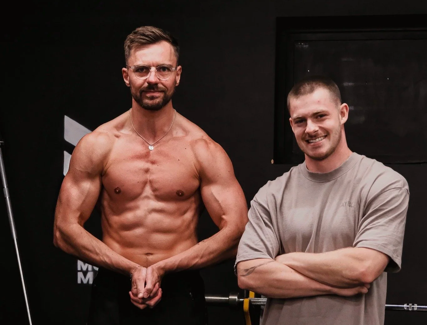 Two men standing indoors against black walls, one shirtless showing muscular physique and the other in a gray T-shirt, smiling with arms crossed.