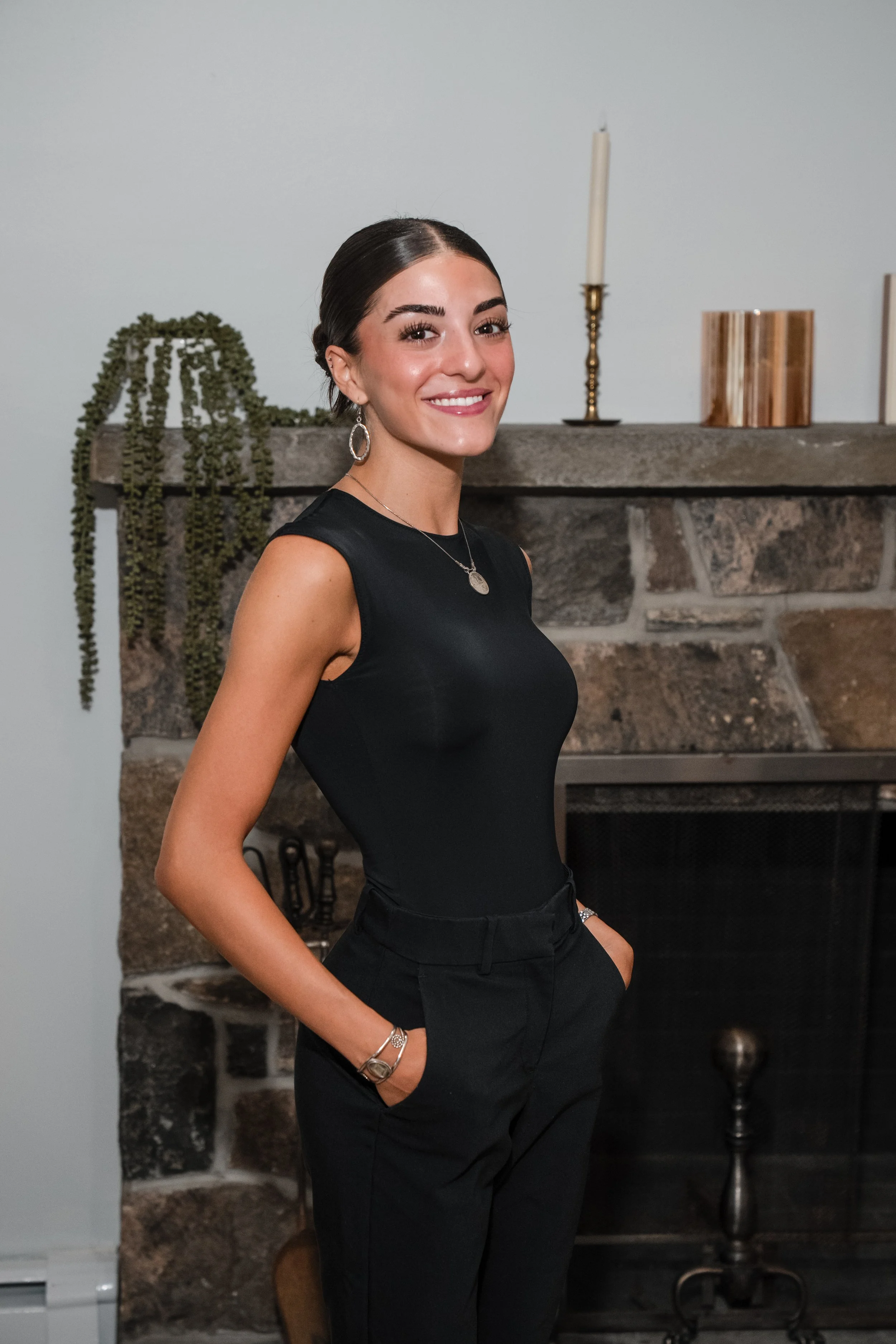 A young woman with dark hair pulled back, smiling, wearing a black sleeveless top, black pants, a necklace, earrings, and a bracelet, standing in front of a stone fireplace with decorative items including a candlestick, a wooden container, and a plant.
