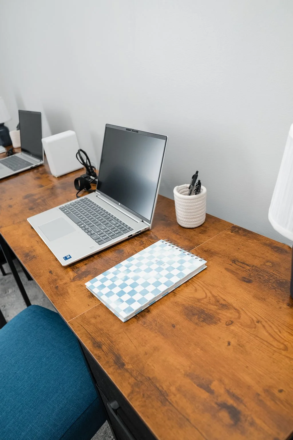 An organized wooden office desk with a silver laptop, a spiral notebook, a white ceramic mug holding pens, and two additional laptops in the background.