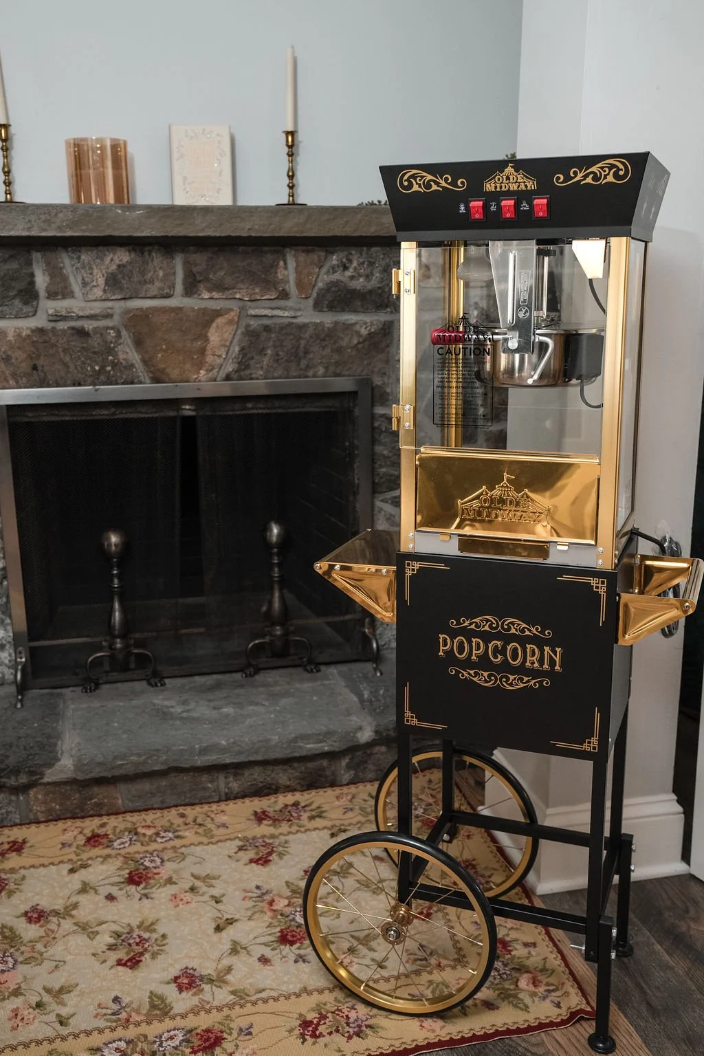 A vintage-style popcorn machine with gold accents and wheels, placed in front of a fireplace with a stone surround and a family photo, on a patterned area rug.