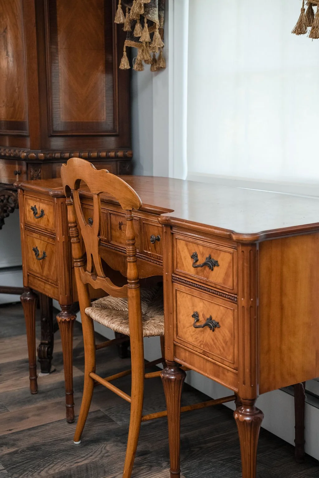 A vintage wooden desk with a light-colored top and intricate drawer handles, accompanied by a matching wooden chair with a woven seat, in a room with wooden flooring and a large white curtain.