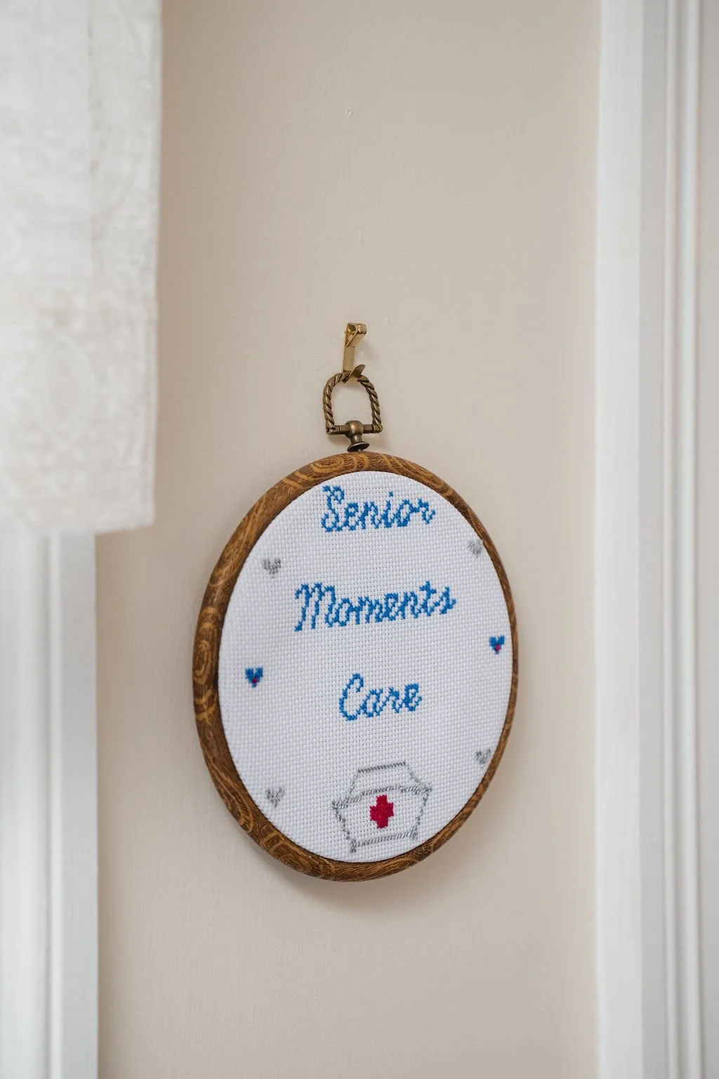 Cross-stitched embroidery hoop with the words "Senior Moments Care" and a small red cross symbol at the bottom, hanging on a wall.