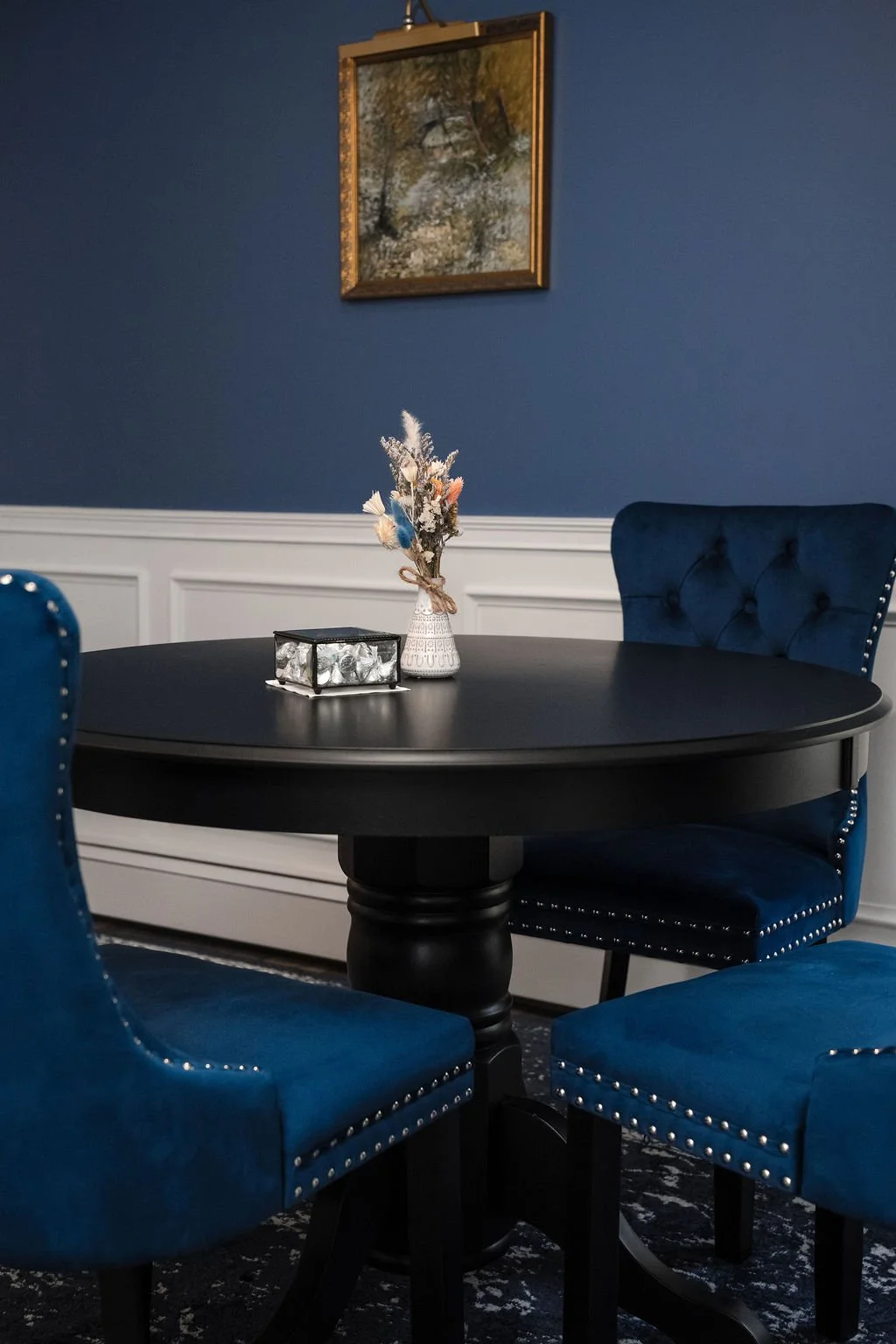 A dark round dining table with a small floral vase and a decorative box in the center. Blue upholstered chairs surround the table. The background has a dark blue wall with a framed landscape painting above the table.