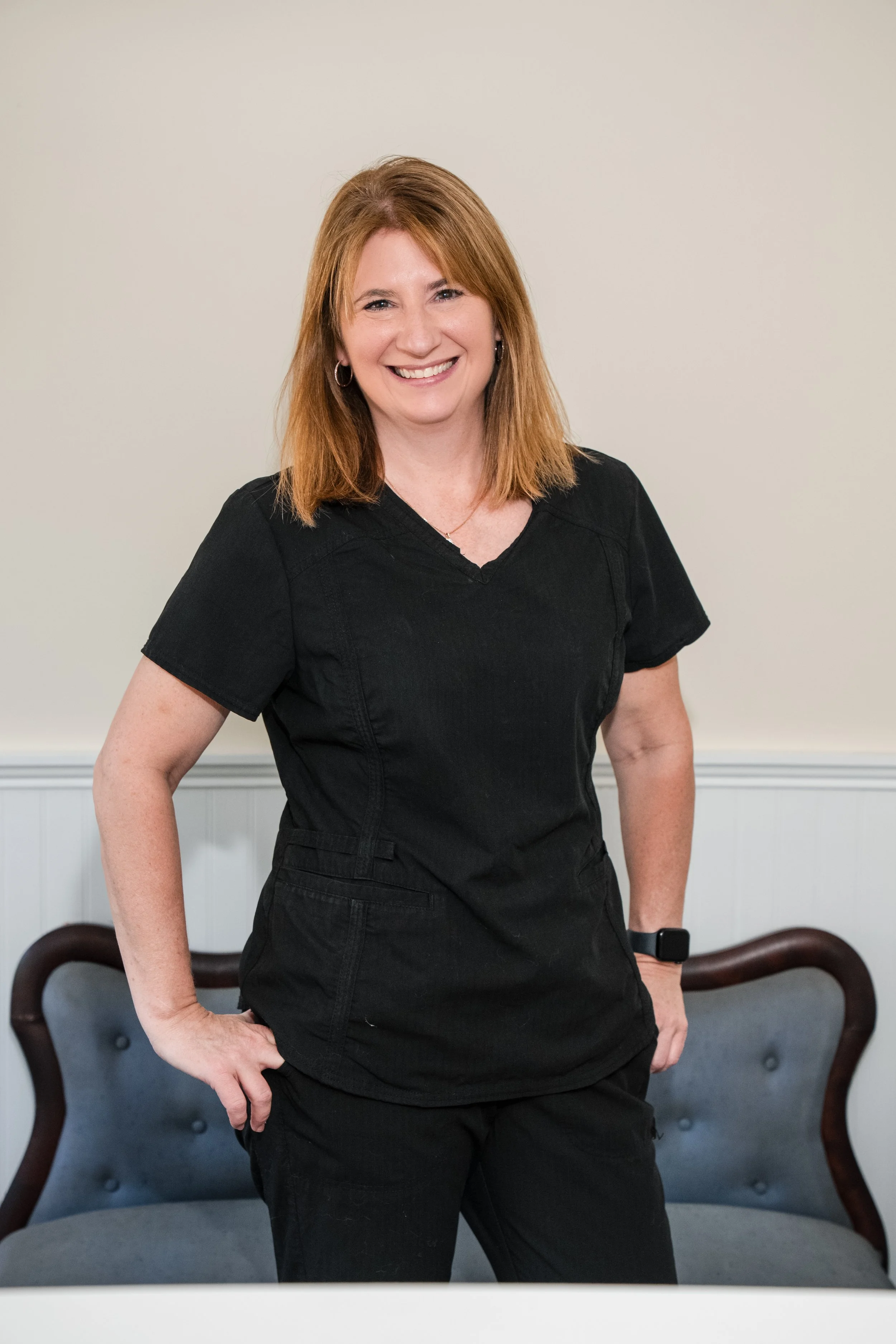 A woman with red hair, wearing a black top and black pants, standing in a room with a beige wall. She is smiling and has an Apple Watch on her left wrist. There are two chairs with dark wooden arms behind her.