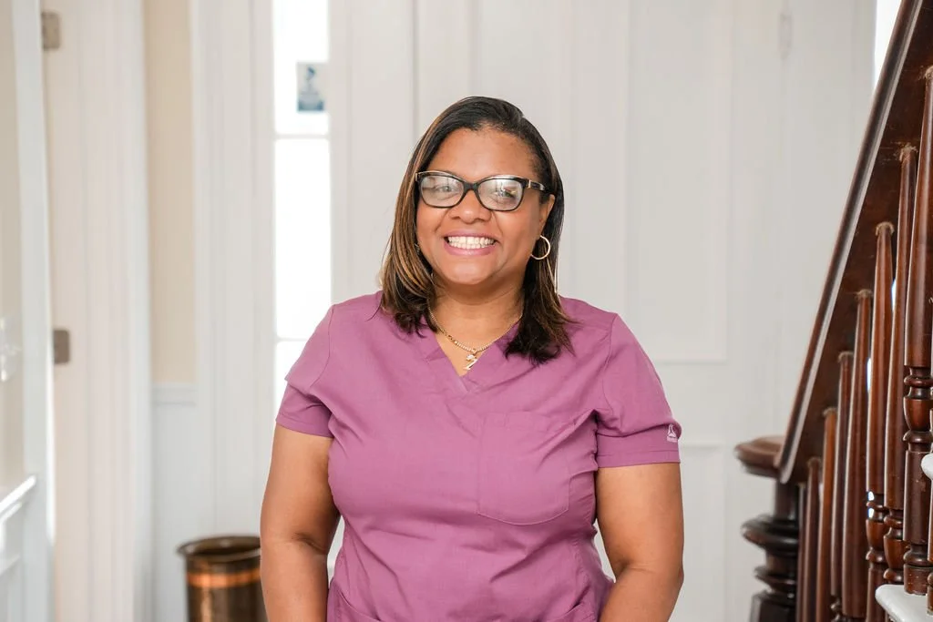A woman wearing glasses and a purple medical scrub top, smiling while standing indoors near a staircase with wooden railing.