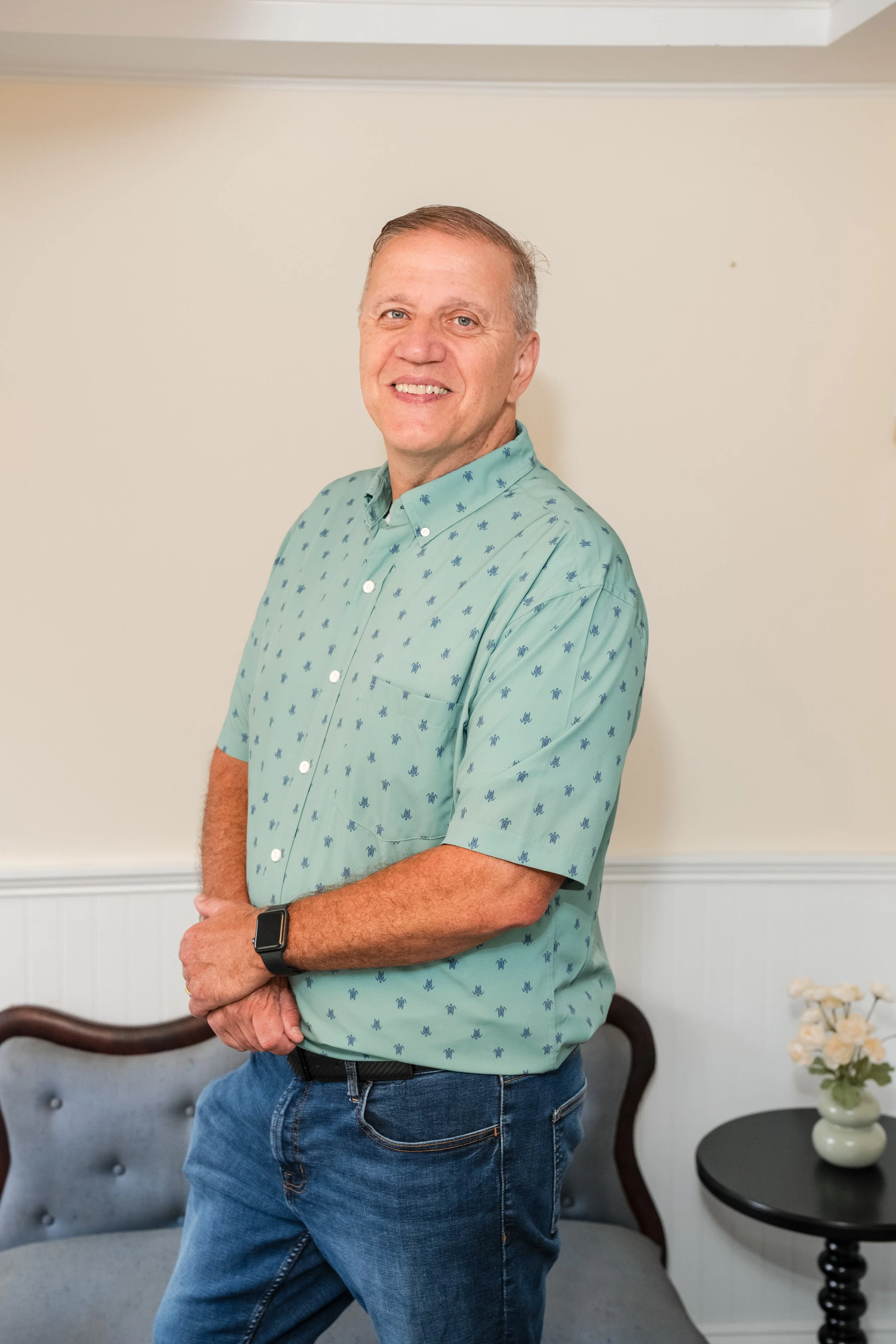 A man with short gray hair smiling, wearing a light blue patterned button-up shirt and blue jeans, standing indoors near a gray tufted couch and a side table with a vase of white flowers.