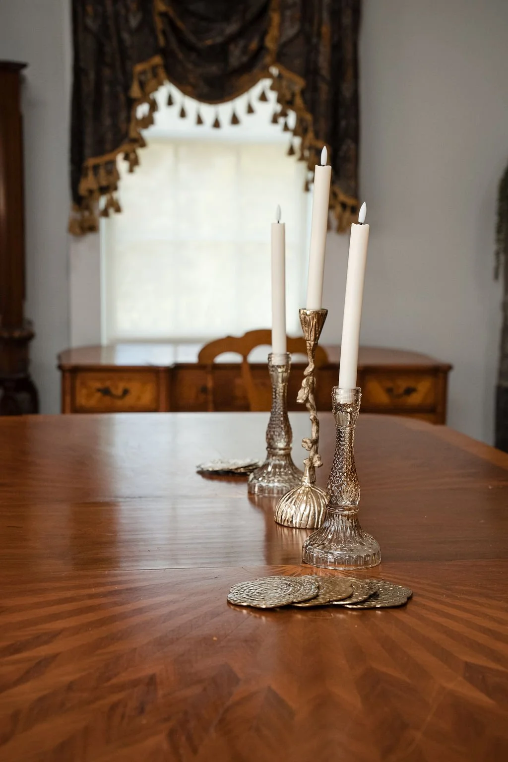 Elegant dining table with three ornate glass candlesticks holding white taper candles, gold decorative coins and a lace doily, in a room with a wooden sideboard, window with valance, and drapery.