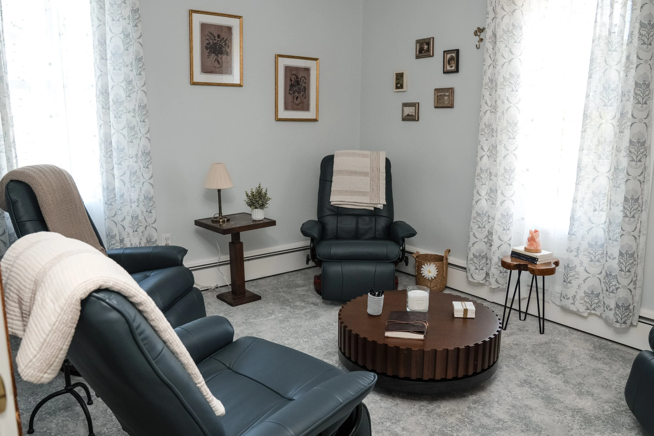 A cozy living room with light blue walls and white floral curtains. Contains black leather chairs, a small wooden side table with a lamp and plant, a round coffee table with books and candles, and framed artwork on the walls.