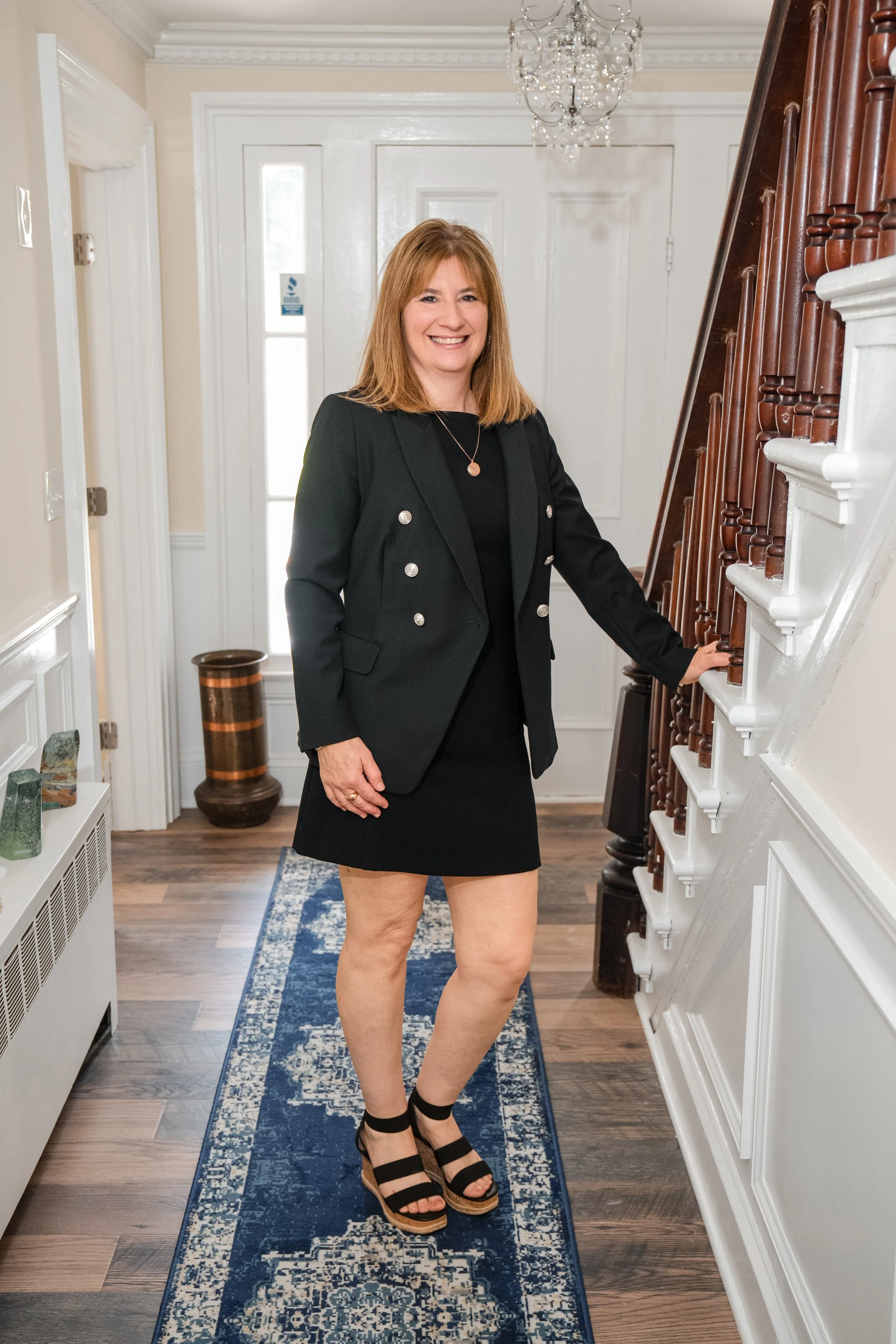 A woman with shoulder-length red hair smiling, wearing a black blazer with silver buttons over a black top and a black skirt, standing on a blue patterned rug in an entryway with hardwood floors, white walls, a chandelier, and a staircase with wooden banisters.